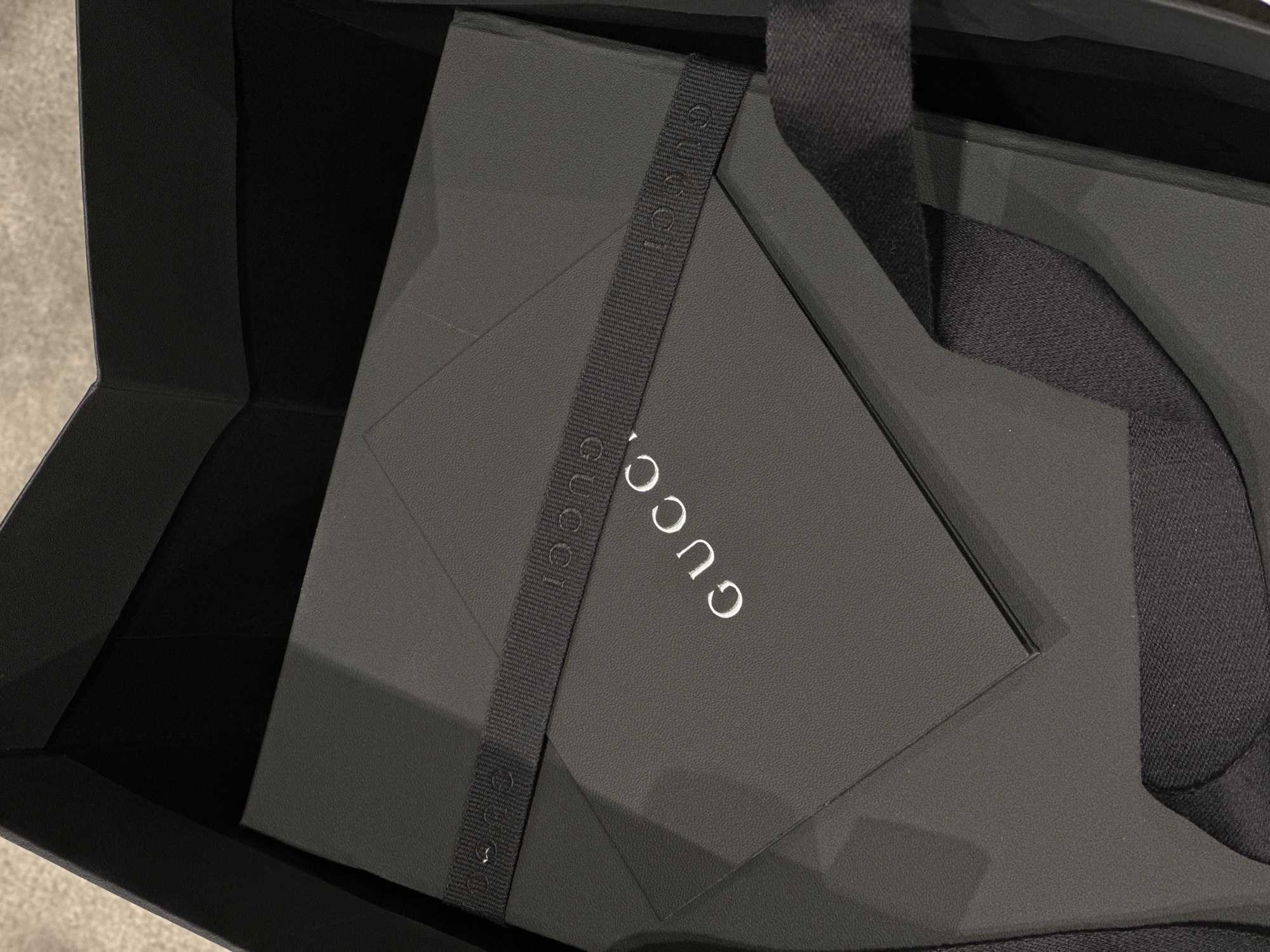 An overhead, close-up shot captures a black luxury box, partially open and nestled within a dark shopping bag or larger presentation container. The box prominently features the brand name "GUCCI" in white capital letters on its lid. A black, textured ribbon, also embossed with repeating "GUCCI" logos, diagonally crosses the top of the box.
The interior of the surrounding bag/container is black, creating a sleek and sophisticated aesthetic. The materials appear to be high-quality, with subtle textures visible on the box and ribbon. The background, visible only slightly on the bottom left, is a light, textured surface, possibly a carpet or floor. The lighting is soft, highlighting the contours and textures of the packaging. The image suggests a moment of unboxing or presentation of a luxury item, likely in an indoor setting, possibly in Paris, France.