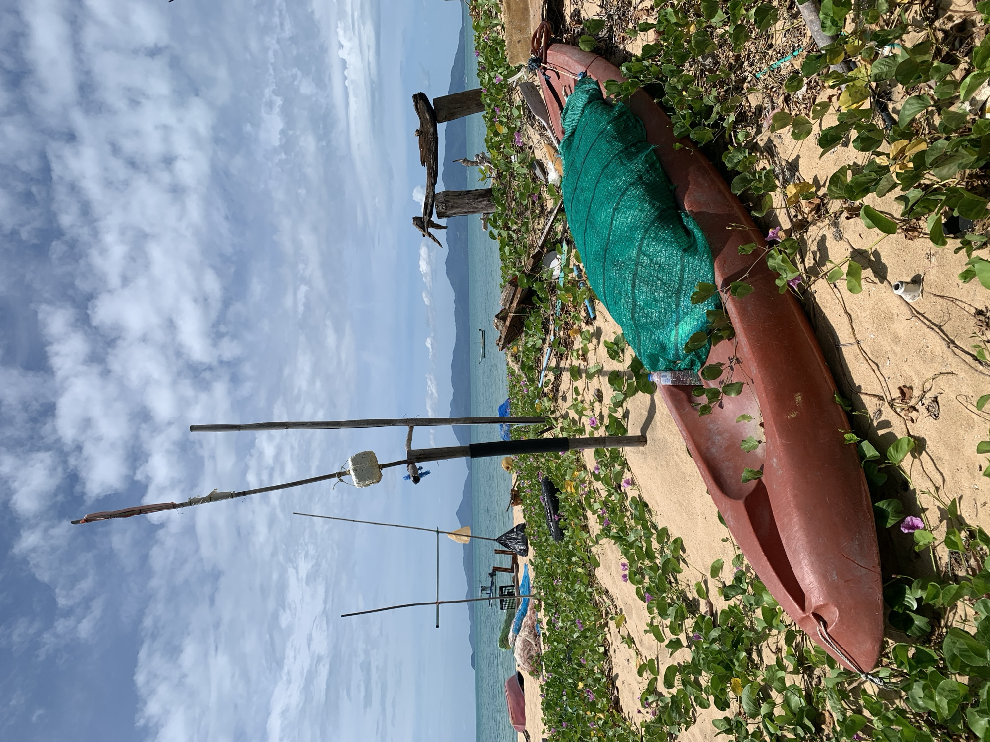 This is a ground-level, eye-level, wide shot of a beach on Ko Samui District, Thailand, with a kayak in the foreground, partially covered by a green net. The kayak is red and lies on the sand, surrounded by green vegetation with small purple flowers. In the middle ground, there are several wooden poles and other debris, including fishing nets and parts of boats. Beyond the beach is the ocean, with a few small boats visible in the distance, and a hazy mountain range under a cloudy sky. The overall scene suggests a somewhat neglected or working beach, possibly used by local fishermen.
