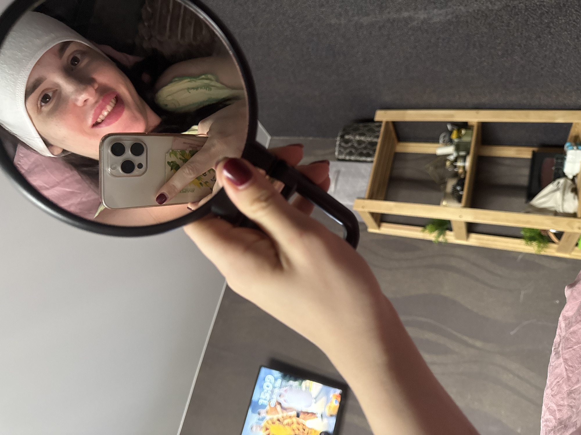A person holds a mirror, reflecting a selfie of themselves. The person in the reflection has a white cap on their head and is smiling. They are holding a smartphone. In the background are various items on a shelf. A separate smartphone is on a surface, displaying the time as 15:09.
