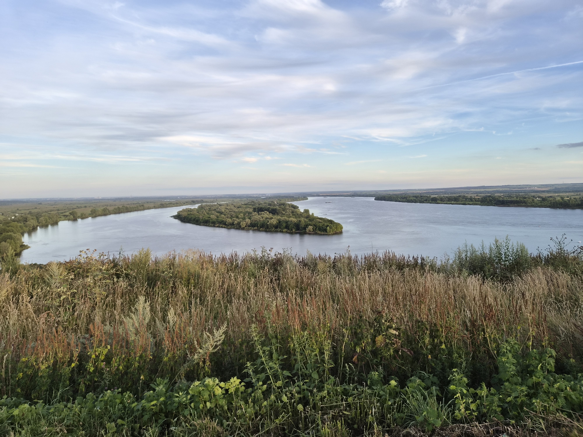 The image captures a panoramic view of the confluence of the Kama and Vyatka rivers in Yelabuga, Russia. The foreground is dominated by tall, dry grasses and green vegetation, suggesting an autumnal landscape. The middle ground features a wide, calm river with a verdant island covered in trees. In the background, the riverbanks are lined with more trees and stretches of flat land, possibly agricultural fields or meadows. The sky above is a mix of blue with scattered, wispy clouds. The overall scene depicts a serene natural landscape.