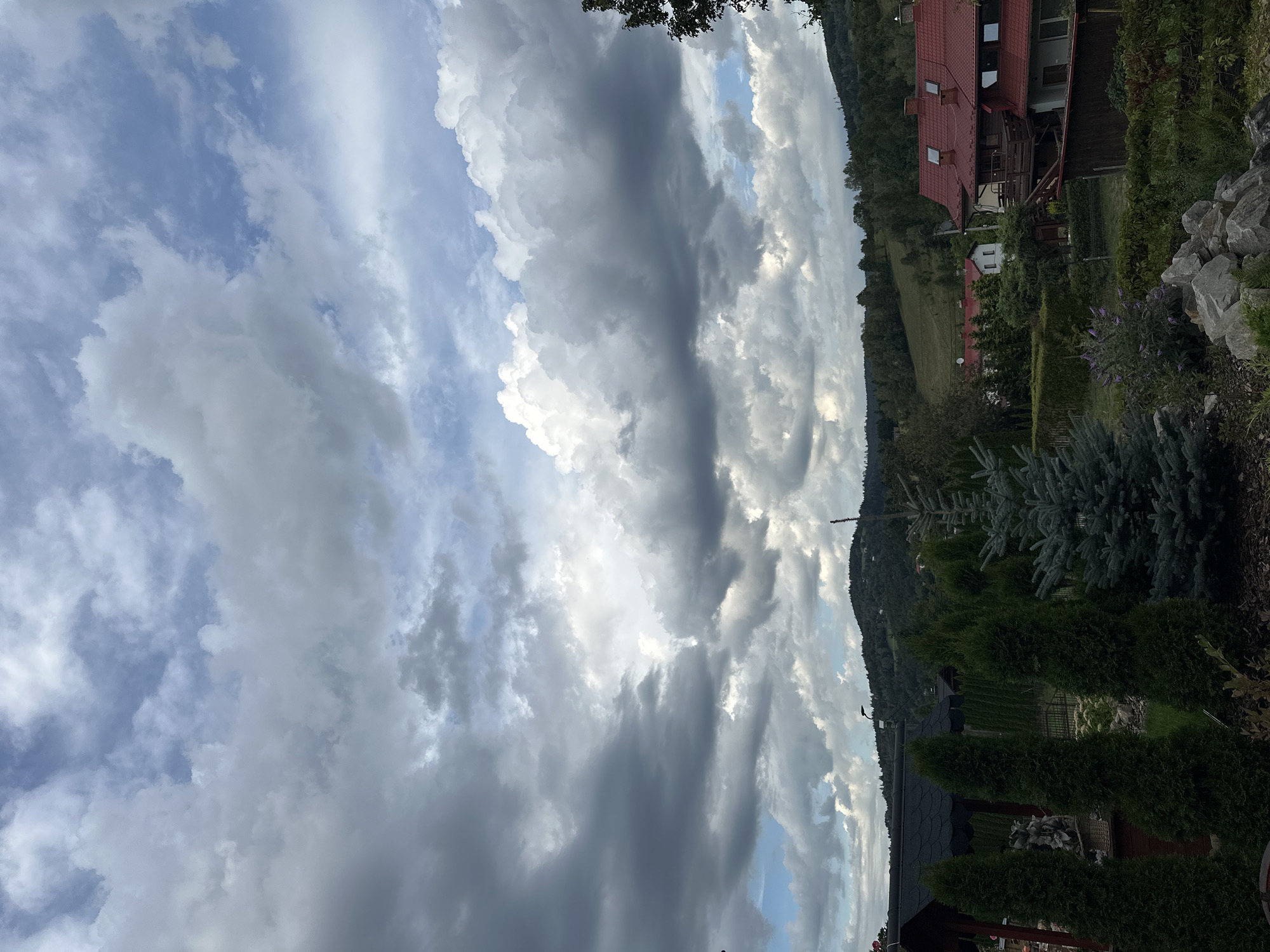The image shows a scenic view of a rural area in Wisła, Poland, under a dramatic sky filled with large, puffy clouds. In the foreground, several coniferous trees are visible, including a tall, slender cypress and a blue spruce. To the left, a dark-roofed structure, possibly a gazebo or an outdoor seating area, is partially obscured by the trees. Behind the trees, a residential area with houses featuring red tiled roofs can be seen nestled amongst the lush green landscape. Rolling hills covered with trees dominate the background, with a hazy blue sky peeking through the clouds. The overall impression is one of natural beauty and tranquility.