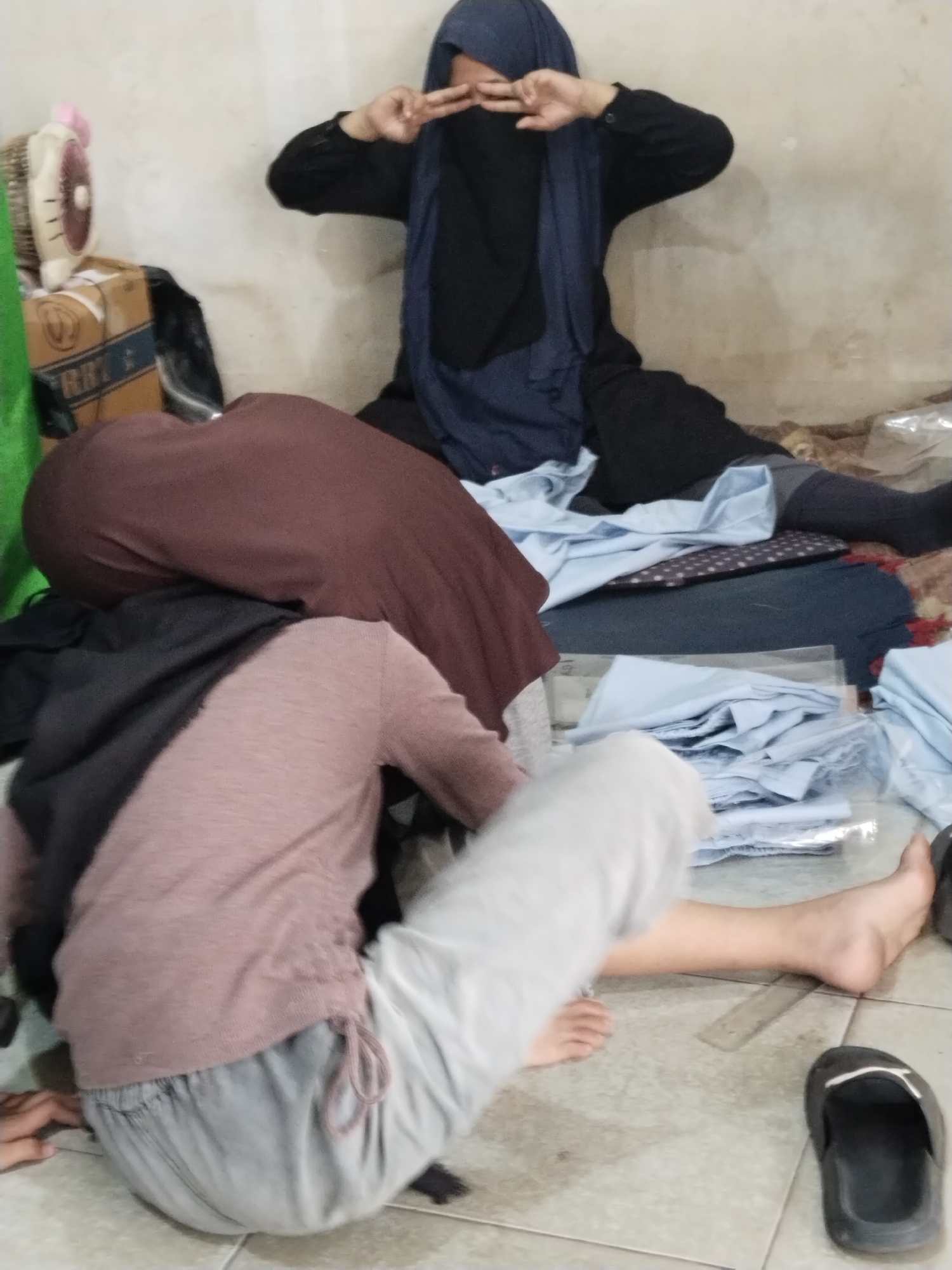 In an indoor environment with a neutral-colored, textured wall and tiled floor, three individuals are visible, two of whom are wearing hijabs and dark clothing. The person in the foreground, positioned centrally, is sitting on the floor with their back to the camera, wearing a pale pink long-sleeved shirt and grey drawstring pants. Their left leg is extended forward, and their right leg is bent, with their right foot visible near a black sandal on the floor. To the left of this individual, another person is partially visible, covered by a dark maroon garment. Behind them, a third person is seated with their legs crossed, wearing a dark blue hijab and a black long-sleeved garment. This individual is making a peace sign with their right hand, positioned near their face. Scattered around the individuals are various items, including fabric, boxes, and a small fan. One box, with visible branding, is to the left of the frame, near the fan. The overall scene suggests a casual or informal gathering in what appears to be a domestic setting.