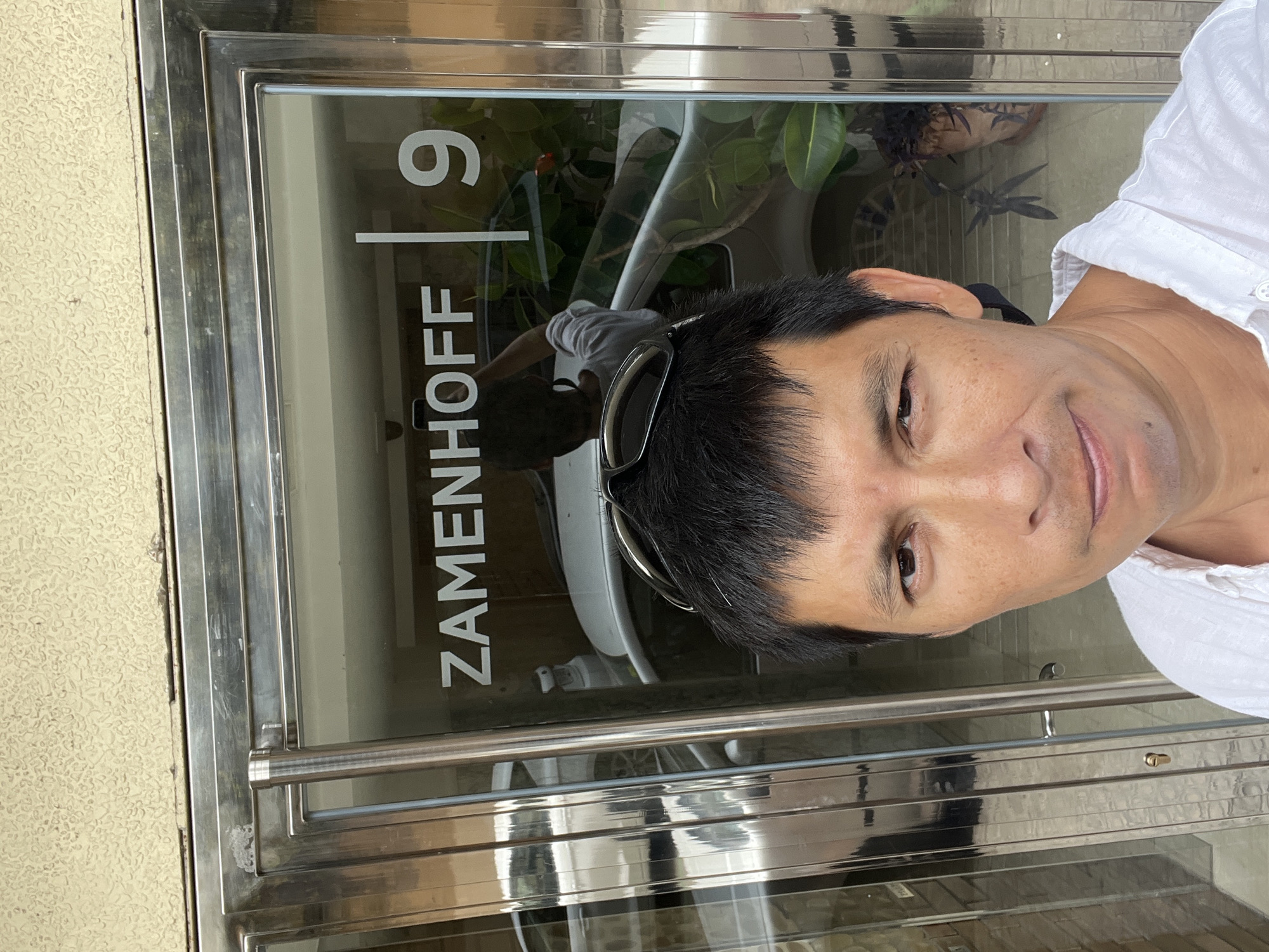 A man is standing in front of a glass door with metal framing. The door has the name "ZAMENHOFF" and the number "9" written on it in white lettering. A white car and some plants are visible through the glass. The man is wearing a white shirt and has sunglasses on his head. The image is a selfie taken at an angle. The background appears to be a building with textured walls. The setting does not provide any specific information to determine the location in Tarragona, Spain.