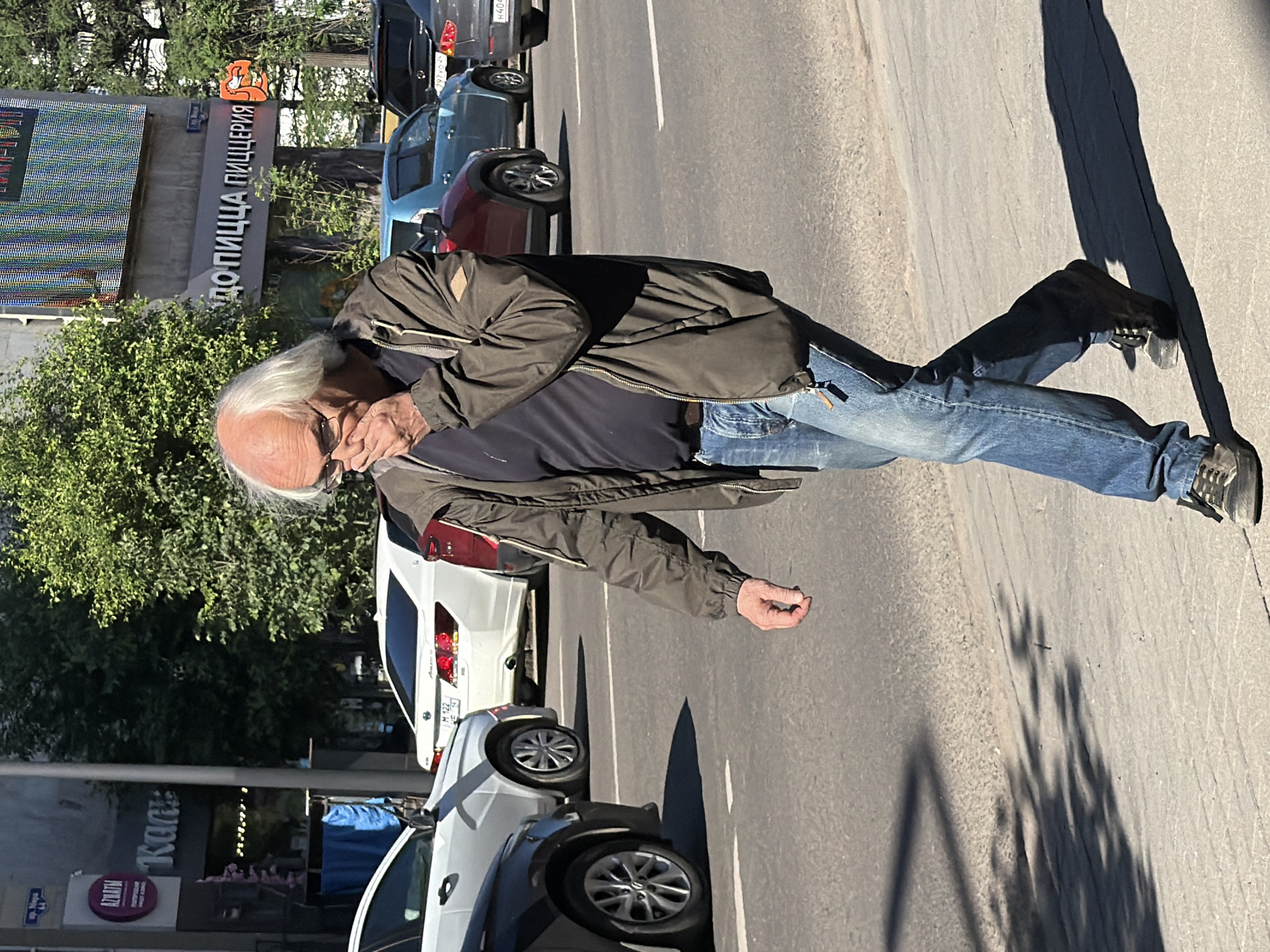 A bald, middle-aged Caucasian man with white hair, wearing glasses, a dark t-shirt, a dark olive green jacket, and blue jeans, walks on a sidewalk next to a street. He is covering his nose and mouth with his right hand. Cars are visible on the street, including a white sedan, a blue SUV, and a dark colored sedan. Buildings are located behind the street, and trees are also present. The scene appears to be in a city environment. Based on the Cyrillic signage visible, specifically "ДОПИЦЦА ПИЦЦЕРИЯ" (Dopizza Pizzeria), which is a chain known to operate in Krasnoyarsk, Russia, and the general architectural style, the location is likely in Krasnoyarsk, Russia. The image was taken during the daytime, as indicated by the sunlight and shadows.