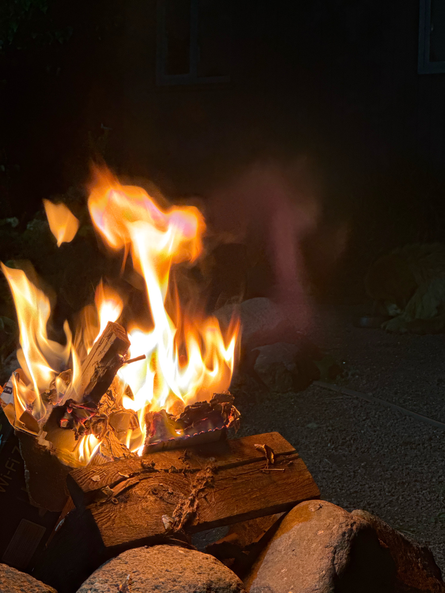 The image depicts a bonfire in an outdoor setting at night. Flames in vibrant oranges and yellows engulf burning wood and paper. Large rocks are visible in the foreground, surrounding the fire pit. In the background, a dimly lit structure with a window is partially visible. To the right of the fire, a hazy, indistinct shape, possibly smoke or a distant object, appears in a purplish hue. Further to the right and slightly obscured by shadow, a light-colored, furry object, possibly an animal, is lying down. The ground appears to be gravel. The scene is consistent with a backyard or a designated outdoor recreational area in Kempelevo, Russia, where a fire might be lit for warmth or ambiance.