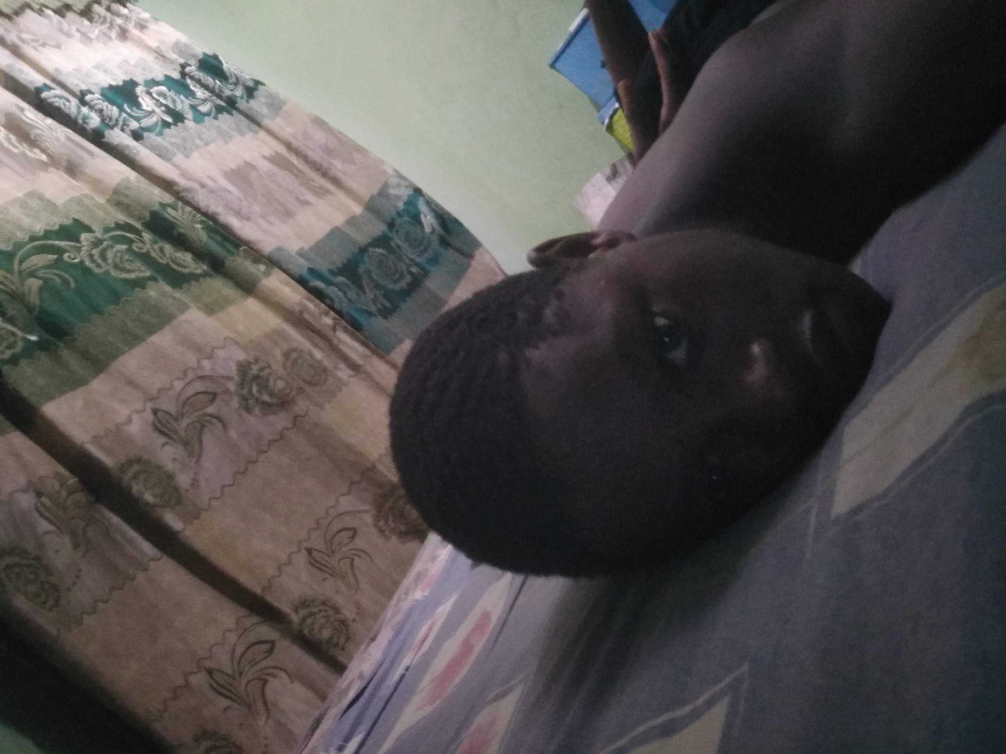 The image depicts a person with dark skin lying on a bed. The person's head is close to the camera and positioned on a pillow with a blue and gray striped pattern. Part of the person's torso is visible. The background includes a patterned curtain in shades of green, beige, and white, and a light green wall. The location is consistent with an indoor setting, potentially a bedroom. The city is Shangisha, Nigeria.