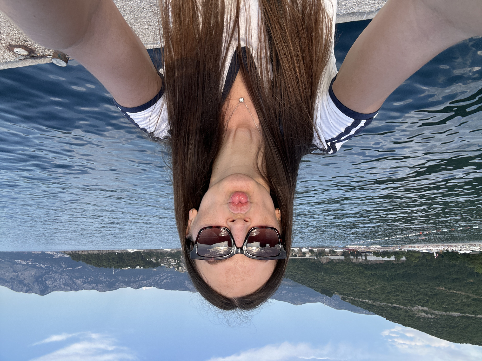 A young woman with long brown hair and sunglasses makes a kissing face as she takes a selfie. She is wearing a white t-shirt with navy blue stripes on the sleeves and a small silver necklace. She stands on a concrete pier with the blue sea in the background. In the distance, a beach with white umbrellas and a lush green mountainside can be seen. The scene appears to be in Budva, Montenegro, likely near a coastal resort.