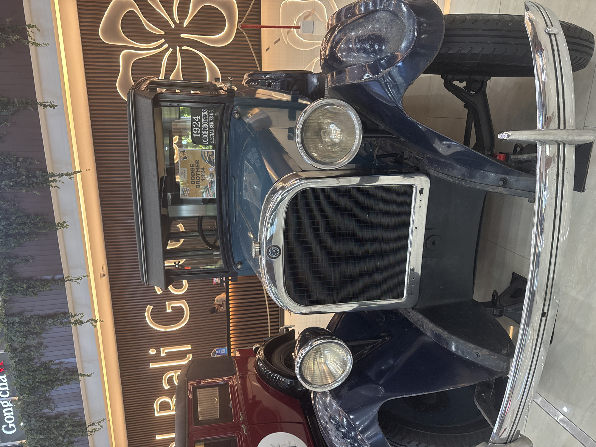 The image shows a close-up view of the front of a vintage blue car, likely a 1924 Dodge Brothers Special Series 116, based on a sign visible in the window. The car is indoors, possibly in a shopping mall or display area. The backdrop features a horizontally striped wall with illuminated floral designs and the inscription "Bali Gal". The foreground also contains green vegetation and the signage "Gong cha". The location appears to be Bali, Indonesia.