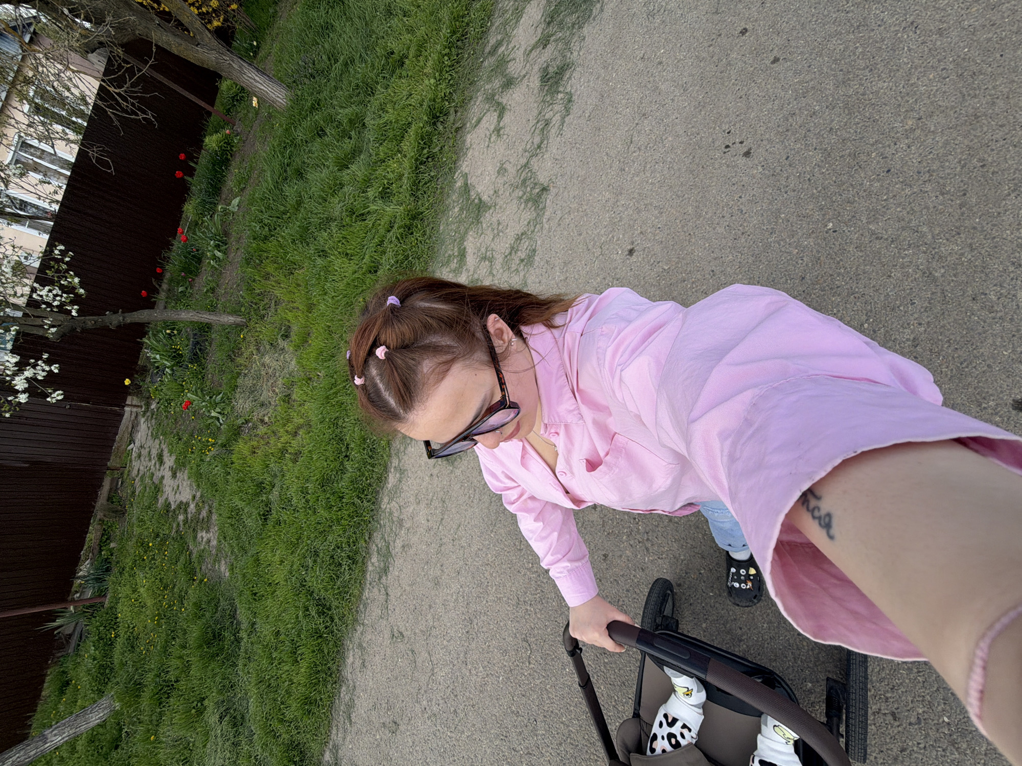 This high-angle, first-person perspective image, taken in Novopokrovskaya, Russia, captures a person walking outdoors while pushing a stroller. The individual, likely a woman, has brown hair styled with small pink and purple clips, and wears black-framed glasses. She is dressed in a light pink button-up shirt and blue jeans. A tattoo in Cyrillic script, possibly "Тилька," is visible on her inner left forearm, and a pink bracelet is on her right wrist.
She is pushing a dark-colored stroller, inside which a baby's feet are visible. The baby wears white socks with black spots (cow print) and yellow smiling faces, along with black shoes adorned with colorful charms. They are on a gray paved path, possibly concrete or asphalt. To the left of the path is a vibrant green grassy area dotted with small yellow flowers and bright red tulips. Beyond the grass, a dark brown fence separates the foreground from a partially visible light-colored building with white-framed windows. Several trees, some with white blossoms, line the area. The scene depicts a calm daytime outing, likely in spring, suggesting a leisurely walk in a park or along a residential path.