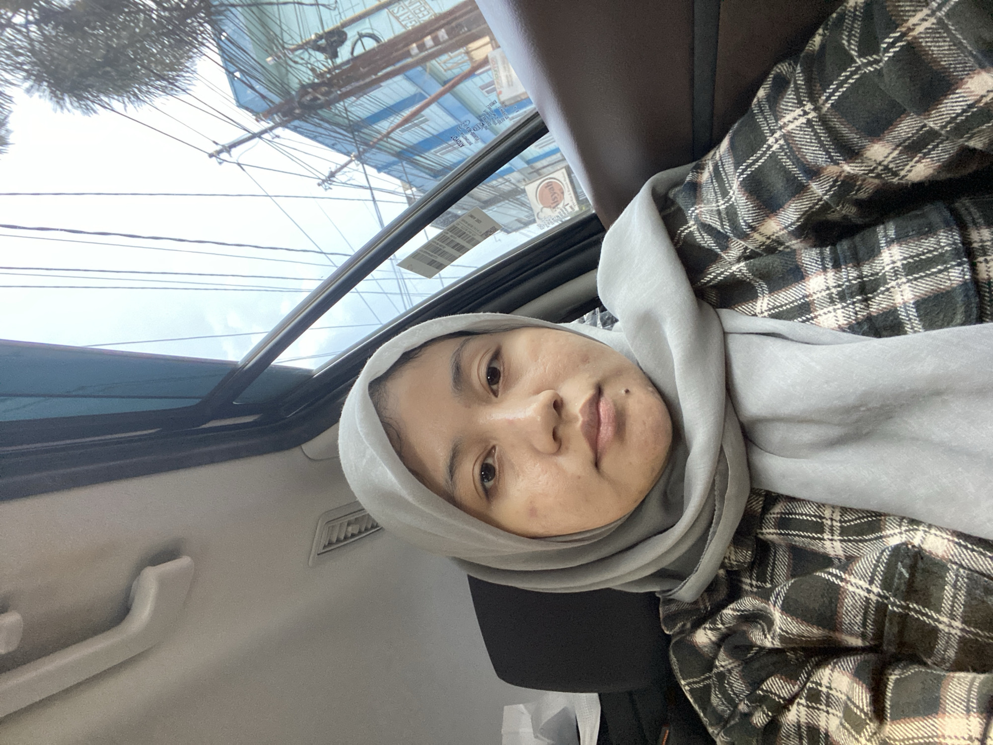 The image is a selfie taken from inside a vehicle, likely a car, with the subject being a woman wearing a light gray hijab and a plaid shirt. She is positioned in the passenger seat, with the interior of the car visible, including a grab handle on the ceiling and the car's door frame. Through the car window, an outdoor urban scene is visible, featuring buildings, utility poles with numerous wires, and trees. The architecture of the buildings suggests a location in Southeast Asia, specifically the city of Banjarmasin, Indonesia, given the signs visible in the background which appear to be in Indonesian. One sign prominently displays "AISYAH". The sky is overcast, and the overall lighting suggests it is daytime.