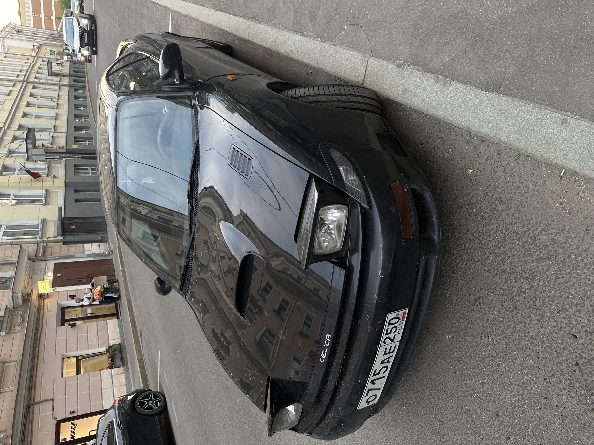 A black 1990s Toyota Celica is parked on a street in Moscow, Russia. The car's license plate is "O715AE250 RUS". The car is parked next to a concrete curb. Buildings are visible across the street. A black car is parked behind the Celica. A light-skinned male on a bicycle is sitting in front of the entrance to a building. Two fair-skinned people are in the building entrance. A building sign for Monique is visible to the bottom left of the image. The image appears to be taken during daylight hours.