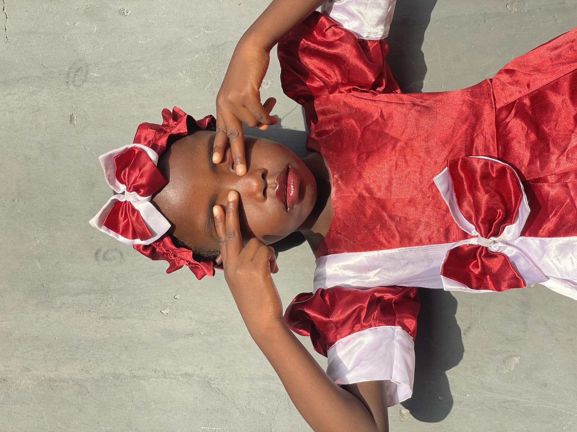 A young child with dark skin is depicted from an overhead view, lying on their back on a textured, light gray concrete or paved surface. The child is wearing a vibrant red and white dress or costume, made of a shiny, satin-like fabric. The dress features ruffled short sleeves with white cuffs and a prominent red and white bow on the waist area. A matching red and white ruffled headpiece adorned with bows frames the child's face.
The child's hands are positioned over their face, with fingers lightly spread across their eyes and nose, creating the impression of peeking through or gently shielding their eyes. Their lips are slightly parted and appear glossy. The scene is illuminated by strong, direct sunlight, casting sharp shadows on the ground and highlighting the child's skin and the sheen of the fabric.
The activity appears to be a playful or contemplative pose outdoors, possibly in bright midday or early afternoon sun, within the context of Badagry, Nigeria. There is no visible text in the image.