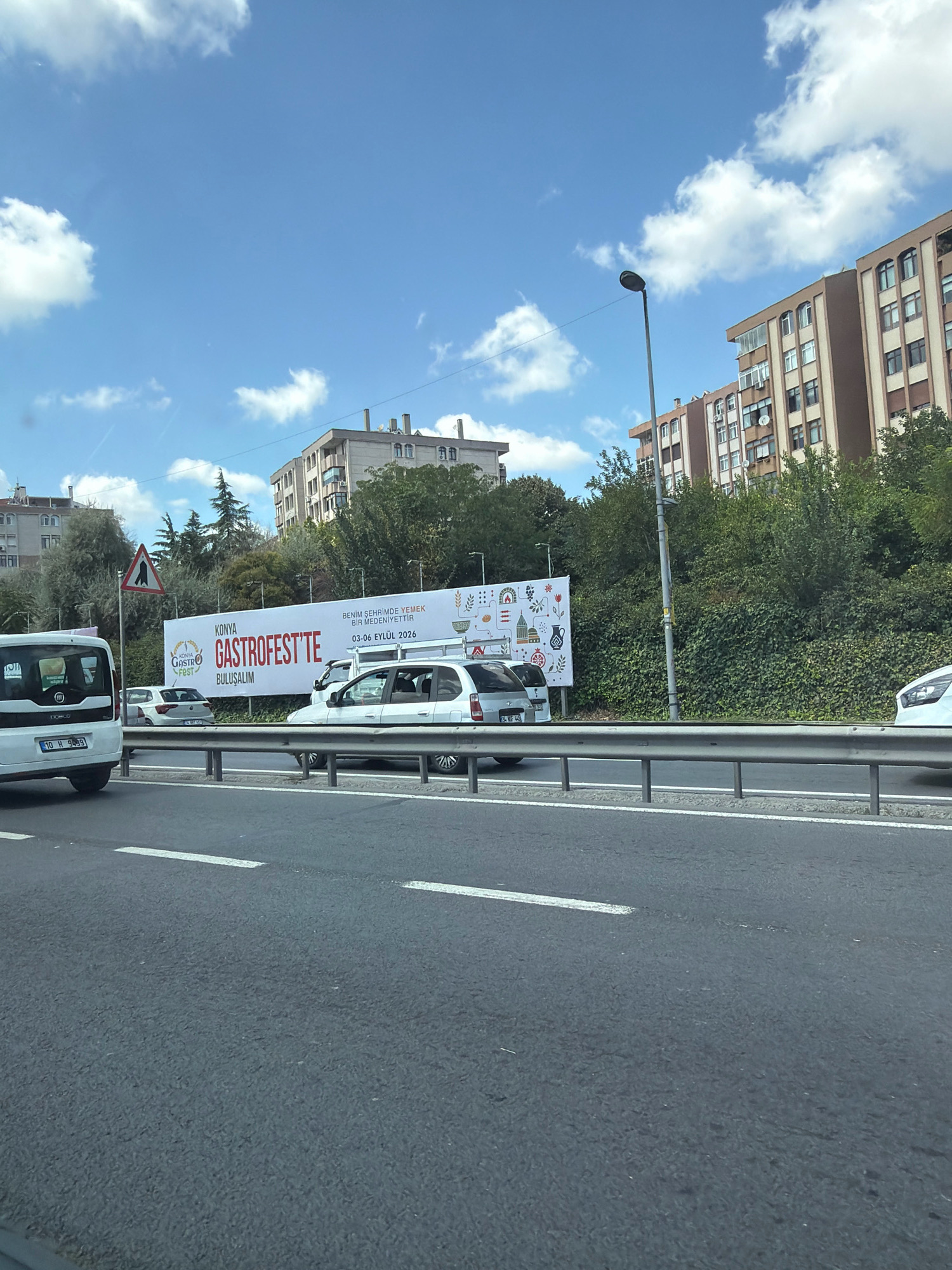 In Bakırköy, Turkey, a large white billboard with red text is visible on the side of a busy road. The billboard advertises the "KONYA GASTROFEST'TE BULUŞALIM" which translates to "Let's meet at Konya Gastrofest". Below this, smaller text reads "BENİM SEHRİMDE YEMEK BİR MEDENİYETTİR" meaning "Food is a civilization in my city". The dates "03-06 EYLÜL 2026" are also displayed. The billboard features a decorative border with illustrations of various food items and historical landmarks. Several cars, including a white van and a silver hatchback, are driving on the road in front of the billboard. The scene is captured on a sunny day with blue skies and scattered white clouds. Apartment buildings and lush green trees form the backdrop to the billboard.