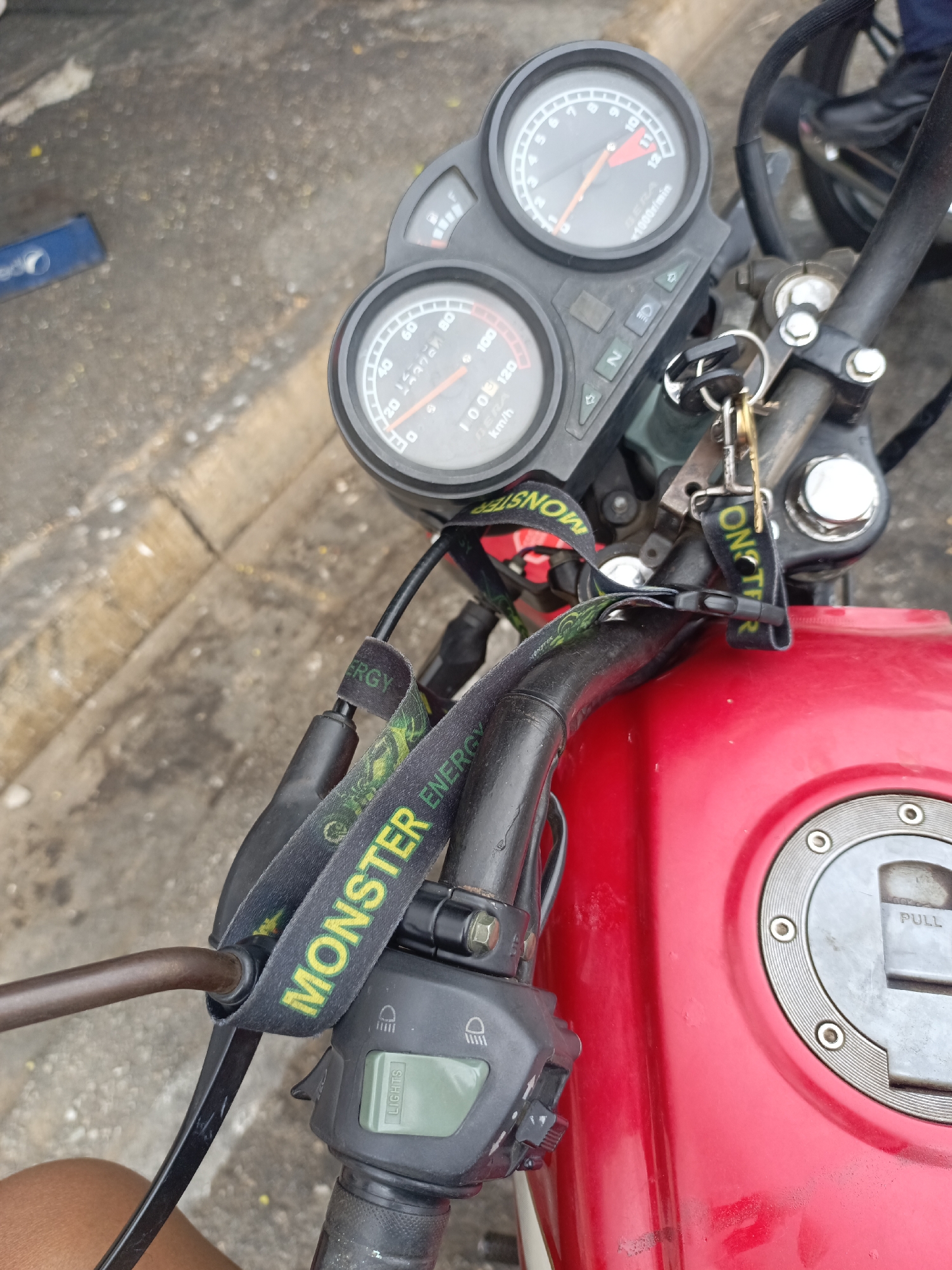 A close-up, high-angle shot reveals the dashboard and handlebars of a red motorcycle. The dashboard features two circular gauges: a speedometer with a reading just under 10 km/h and a tachometer with the needle pointing near 1000 r/min. Keys hang from the ignition. A black lanyard with "MONSTER ENERGY" printed in yellow and green is wrapped around the handlebars. The fuel tank is red with a silver fuel cap labeled "PULL." To the left of the tank are the motorcycle's controls, including a light switch. Based on the OCR data, the gauges are labelled as "BERA". The scene appears to be in an outdoor setting, with the edge of a curb visible in the background. The location is unspecified.