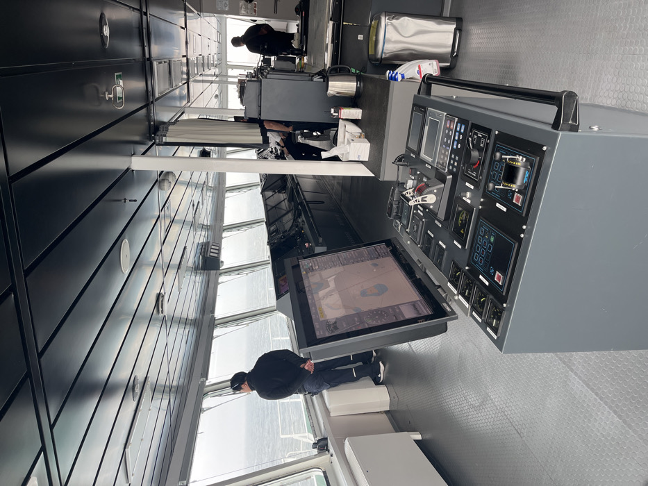 Inside a vessel's bridge, a man in a black jacket and cap stands near a large navigational display screen showing a map. Behind him, a panel filled with various gauges and controls. Other personnel are present further in the room. The interior is modern, featuring dark gray walls, a light gray floor with a textured pattern, and large windows.