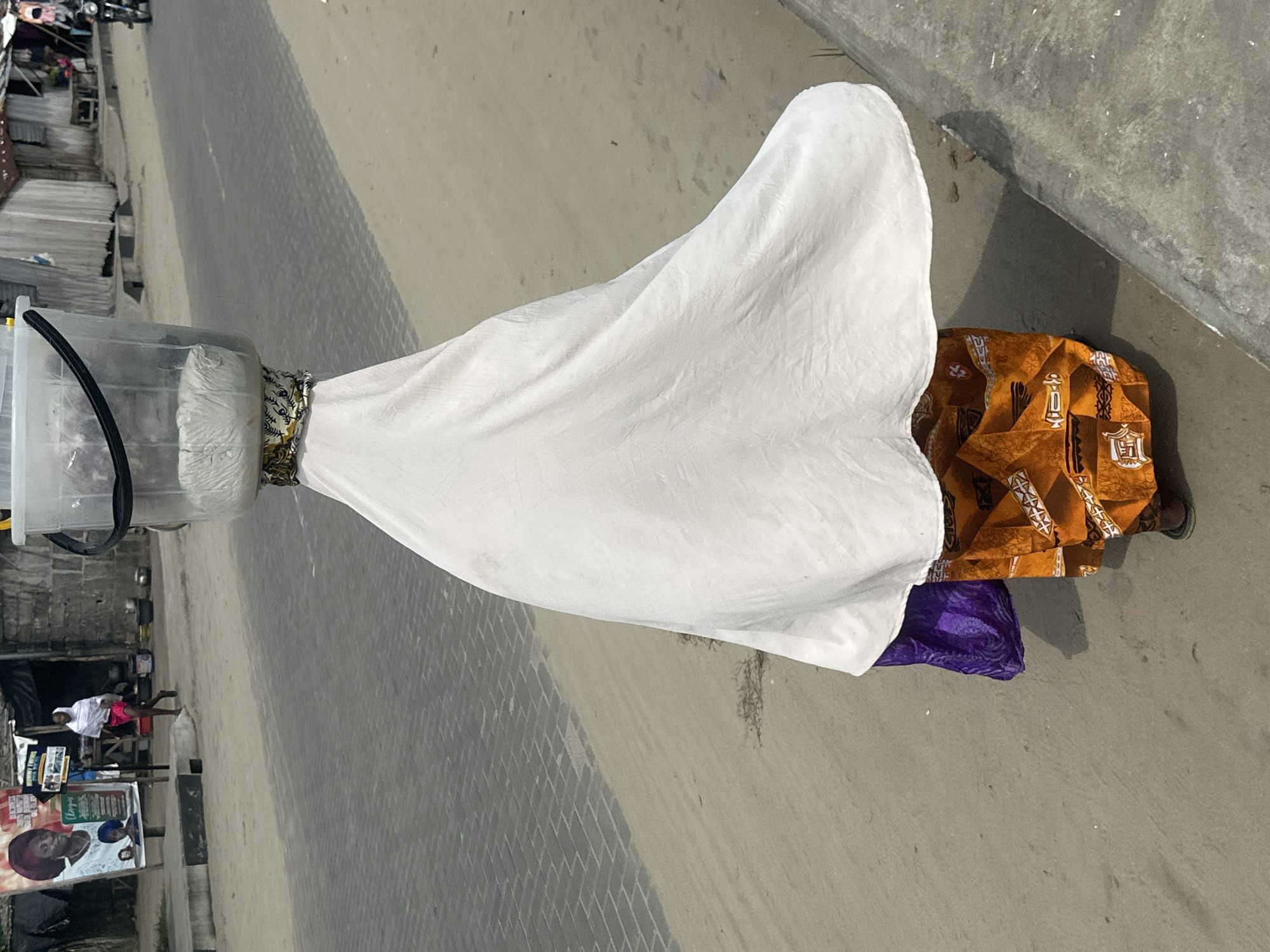 A person is walking away from the camera on a sandy path next to a paved road in Badagry, Nigeria. The person is carrying a large, clear plastic bucket filled with white material on their head, balanced by a patterned cloth wrapped around their head and neck. They are wearing a white cloth draped over their body, a purple fabric underneath, and a vibrant orange and yellow patterned skirt. The background shows buildings, some with corrugated metal roofs, and a poster on a pole. A motorcycle is visible in the distance on the road. The ground is a mixture of sand and paved interlocking bricks.