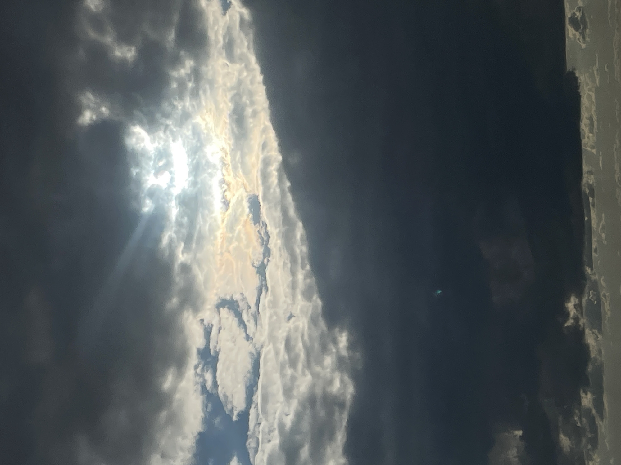 The image, captured in Votkinsk, Russia, displays a dramatic sky scene dominated by extensive cloud formations. The picture is oriented vertically, showing a dynamic interplay of light and shadow across the sky.
The upper left and central part of the frame feature a brilliant burst of sunlight, partially obscured by bright, white, and golden-hued clouds. Sunbeams radiate from this intense light source, illuminating the surrounding cloud edges and creating a radiant effect. Contrasting sharply with this brightness, the right and lower sections of the image are filled with dark, heavy, deep grey clouds, suggesting dense cover or a stormy atmosphere. The overall impression is of a powerful, changing sky, with the sun breaking through an otherwise overcast or turbulent cloud layer.
In the far right edge of the image, a narrow, light-colored vertical structure is visible, possibly part of a building or a pole, indicating the view is from an area with some architectural presence. A small, faint greenish-blue speck, likely a lens flare, is present in the darker cloud area on the right. The scene depicts daytime conditions. No visible text is present in the image.