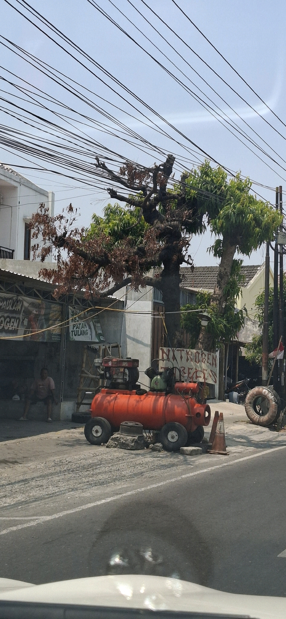 The image depicts a street scene in Colomadu, Indonesia, characterized by a bright, clear sky and a dense network of overhead power lines crisscrossing the frame. In the foreground, the asphalt road surface shows tire tracks and markings. To the right, a white car hood is visible at the bottom edge.
Dominating the mid-ground is a striking, bright orange, wheeled air compressor. It is a large, cylindrical tank with two wheels on each side, appearing to be made from a repurposed barrel. On top of the tank sits a red engine and a green tank, suggesting its operational capacity. The compressor is positioned on a rough, dusty surface, with a concrete block and rocks supporting its base. A small orange traffic cone is placed nearby.
Behind the air compressor, a building with a gray, corrugated metal roof and a white facade is visible. A banner with Indonesian text, which appears to advertise services, hangs on the front of the building. A person, partially obscured, is seated on a low bench or step inside the open doorway of this building. Next to the building, a ladder leans against it.
Further behind these elements, a large, mature tree with a thick, gnarled trunk stands prominently. The tree exhibits a mix of healthy green foliage and dry, brown, withered leaves, indicating seasonal changes or stress. Several other trees with green leaves are also present.
To the right of the air compressor, a spare tire is mounted vertically against a pole. Further down the street, glimpses of other buildings and a parked motorcycle suggest a commercial or residential area. An Indonesian flag is visible on a pole near the tire. The overall impression is a functional, perhaps slightly cluttered, outdoor workspace or roadside service area.