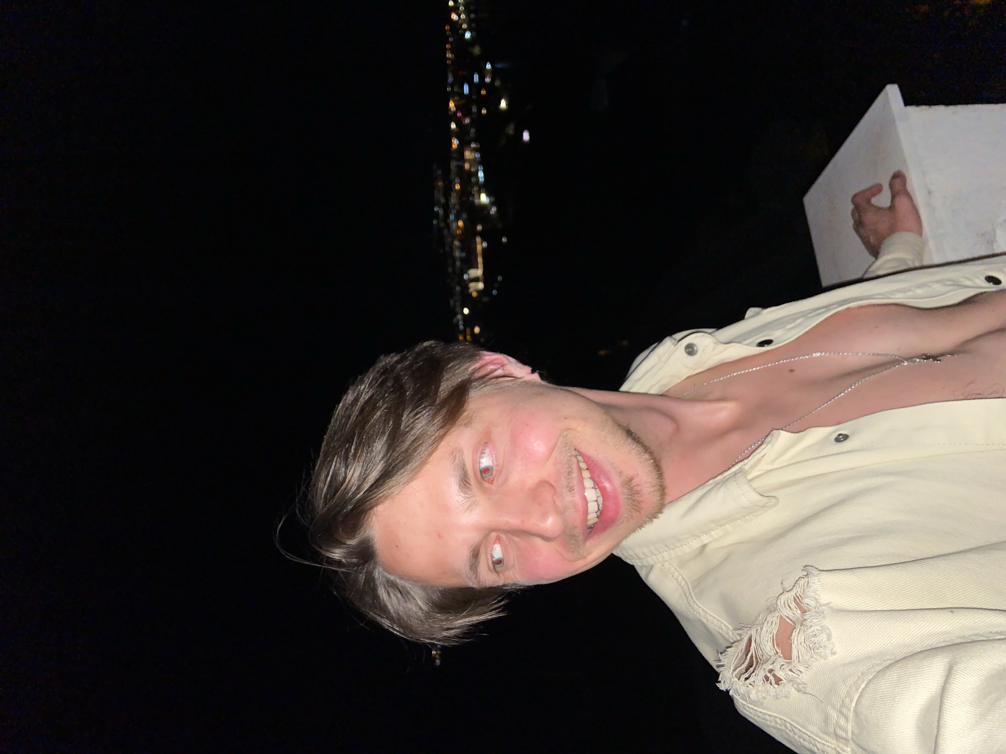 A man with light brown hair, blue eyes, and fair skin is smiling, and looking at the camera. He is wearing a light-colored, button-down jacket, with a silver necklace. To the upper left of the frame, there is an indiscernible grouping of lights that appear to be from the city of Šušanj, Montenegro. The background of the image is black. The man is holding on to a white, square ledge with one hand.