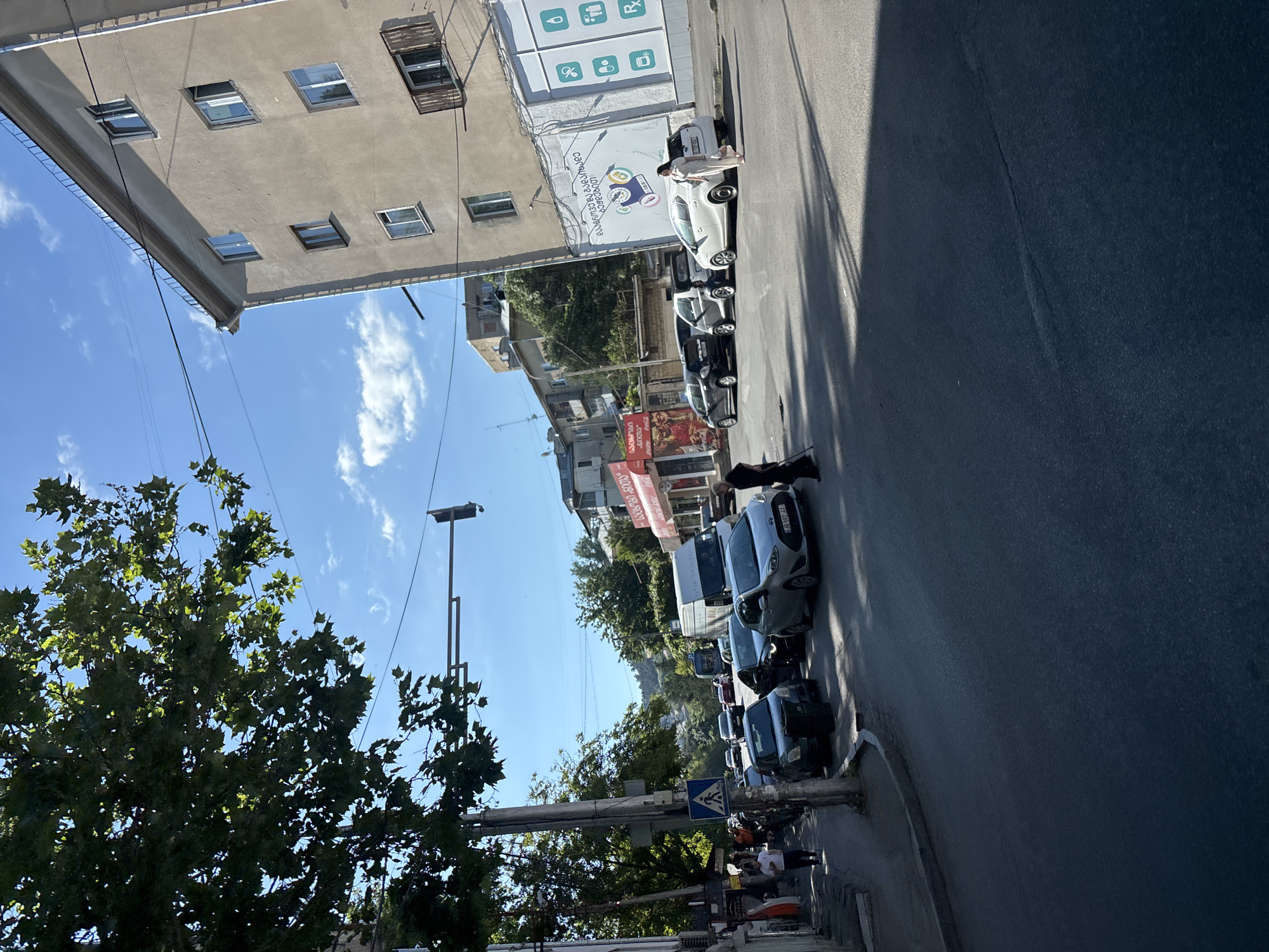 The image shows a street scene in Tbilisi, Georgia, with a traffic light and several cars on a sunny day. A woman in a light-colored dress is walking on the sidewalk next to a white car. In the background, there is a multi-story building with several windows. The street is paved, and there are trees on the left side of the image. The sky is blue with some clouds.