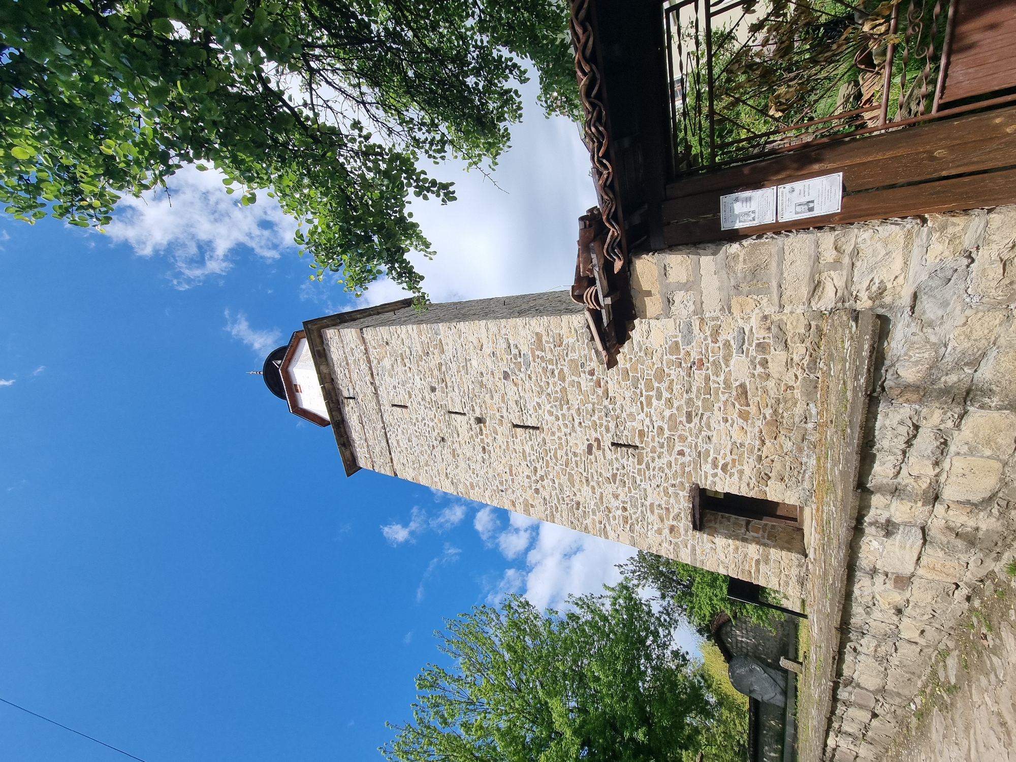 The image shows the Clock Tower in Elena, Bulgaria, viewed from below. The stone tower is rectangular, with small, rectangular openings along its height. It is surrounded by green trees against a blue sky with scattered clouds. A wooden fence or gate is visible on the right, attached to a stone wall. Two posters are affixed to the wooden part of the structure. The tower and fence are constructed of stones of various sizes. The photo seems to be taken at a tilted angle, looking up at the tower.