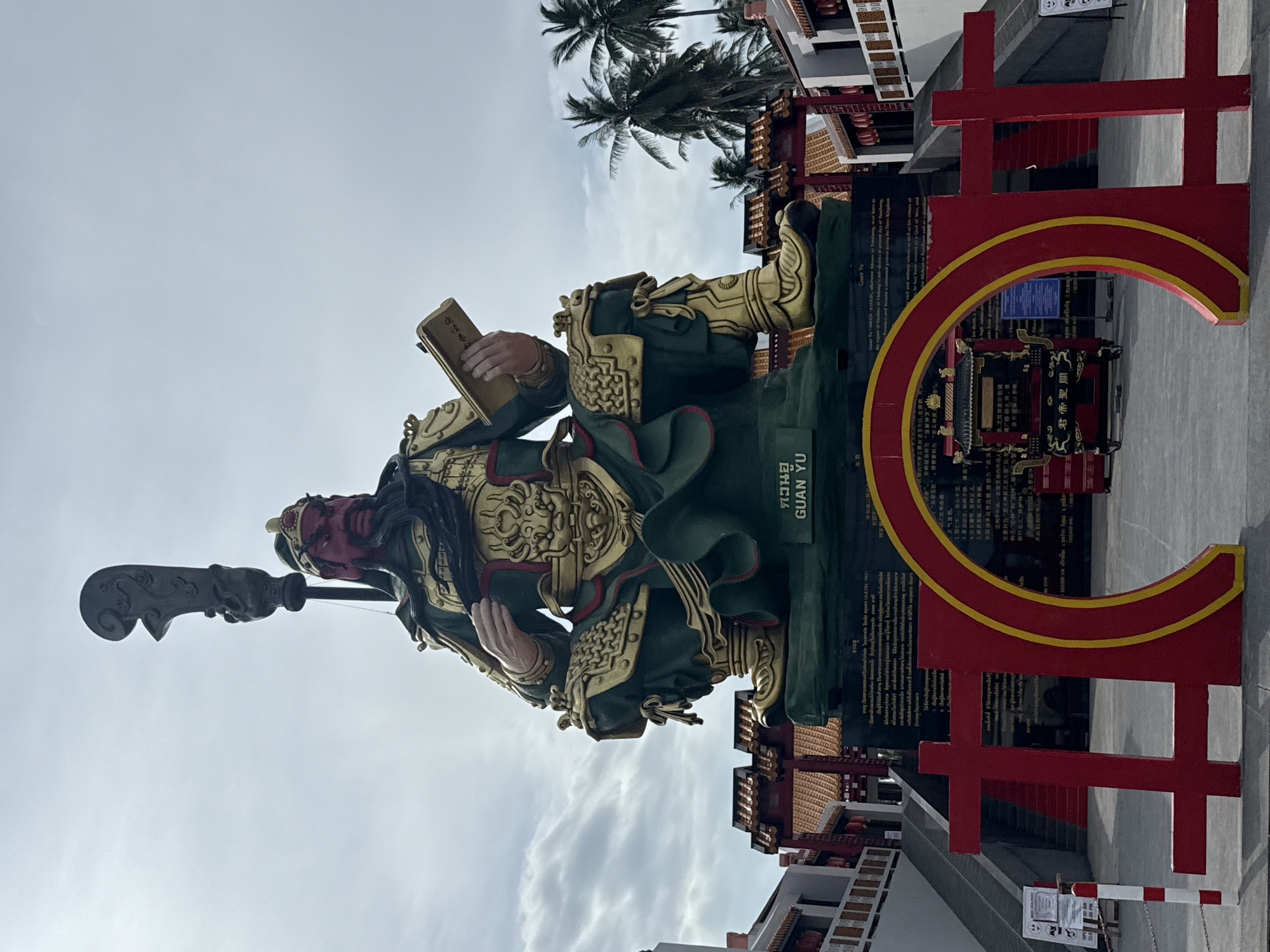 A large, imposing statue of Guan Yu, a Chinese deity and historical figure, sits on a raised platform in Ko Samui District, Thailand. The statue is predominantly green and gold, depicting Guan Yu in full armor, seated with a book in his left hand and a large weapon, a guandao, held aloft in his right. The statue is positioned in front of a building with traditional Chinese architectural elements, including red roofs and ornate decorations. In the foreground, a large red and yellow archway frames the view of the statue and the surrounding structures. Palm trees are visible in the background, suggesting a tropical setting. The sky is overcast with clouds. The base of the statue features a black plaque with text in both Thai and Chinese, identifying the deity and providing some historical context. A small, ornate altar or shrine is visible behind the red archway.