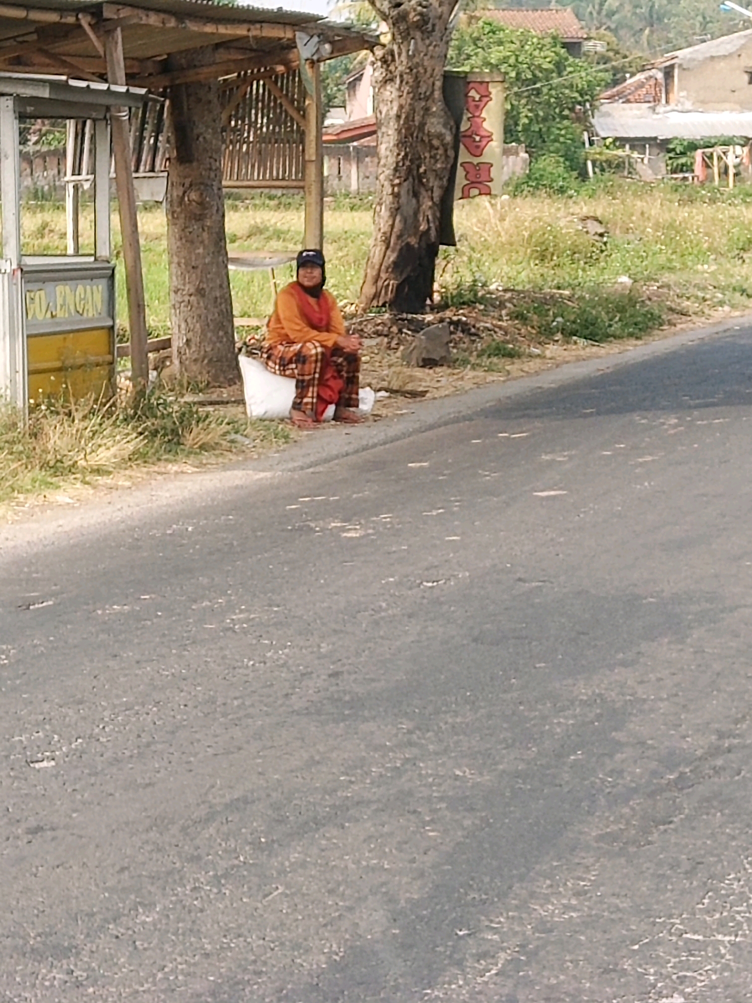 A woman sits on a white object next to a roadside structure in Garut, Indonesia. She is wearing a long-sleeved orange shirt, a patterned headscarf, and a plaid skirt. She is also wearing a dark baseball cap. The structure appears to be a small, open-air shelter with a corrugated metal roof and wooden supports. In front of the shelter, there is a white and yellow kiosk. Behind the woman, a tree trunk stands prominently, and further in the background, there are houses with red tile roofs and lush green vegetation. The foreground is dominated by an asphalt road, with some scattered debris. The overall scene suggests a rural or semi-rural setting.