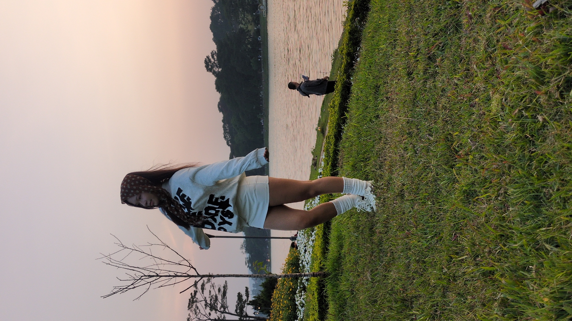 A young woman with long dark hair is seen standing on a grassy area near a lake in Da Lat, Vietnam. She is wearing a light-colored hoodie with dark lettering, light-colored shorts, white socks, and white sneakers. A patterned scarf is draped over her head.
In the background, the lake is visible with trees along its edge. A person can be seen standing at the water's edge. The sky is a pale pinkish-grey color, suggesting it is either dawn or dusk. A bare tree stands near the woman. There are flowers and landscaping around the grassy area. The grass appears to be well-maintained.