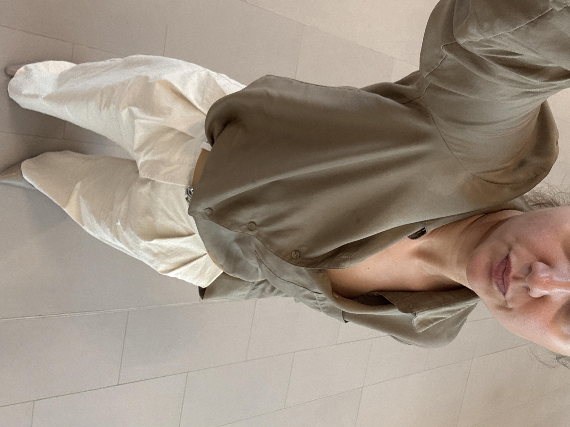 A person wearing beige pants and an olive-green button-down shirt stands indoors on a light-colored tiled floor. The person's face, neck, and upper chest are visible. The scene appears to be a selfie taken indoors, possibly in a residential or commercial building. There's no evidence to suggest the location is in Dubai, United Arab Emirates.