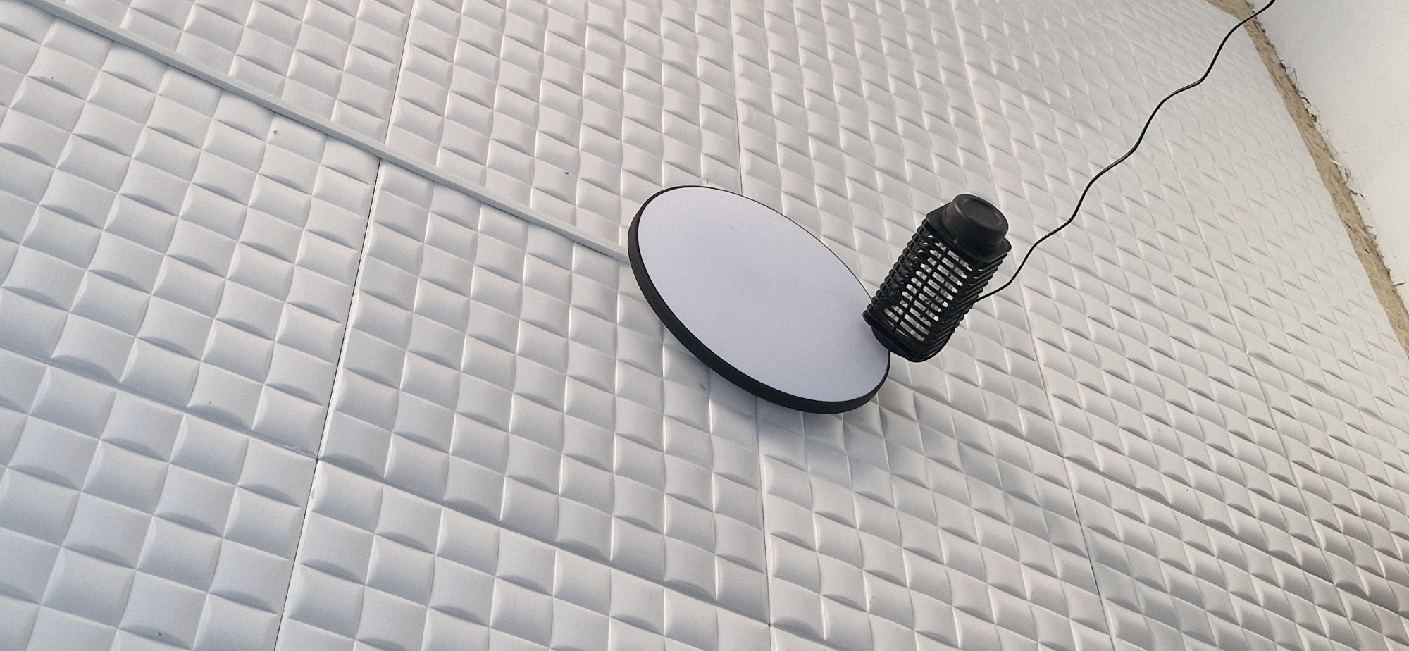 A black mosquito repellent device with a wire is positioned in front of a circular white light fixture, both mounted on a textured white wall. The wall features a repeating pattern of raised diamond shapes. The image captures a ceiling or wall in an interior space.