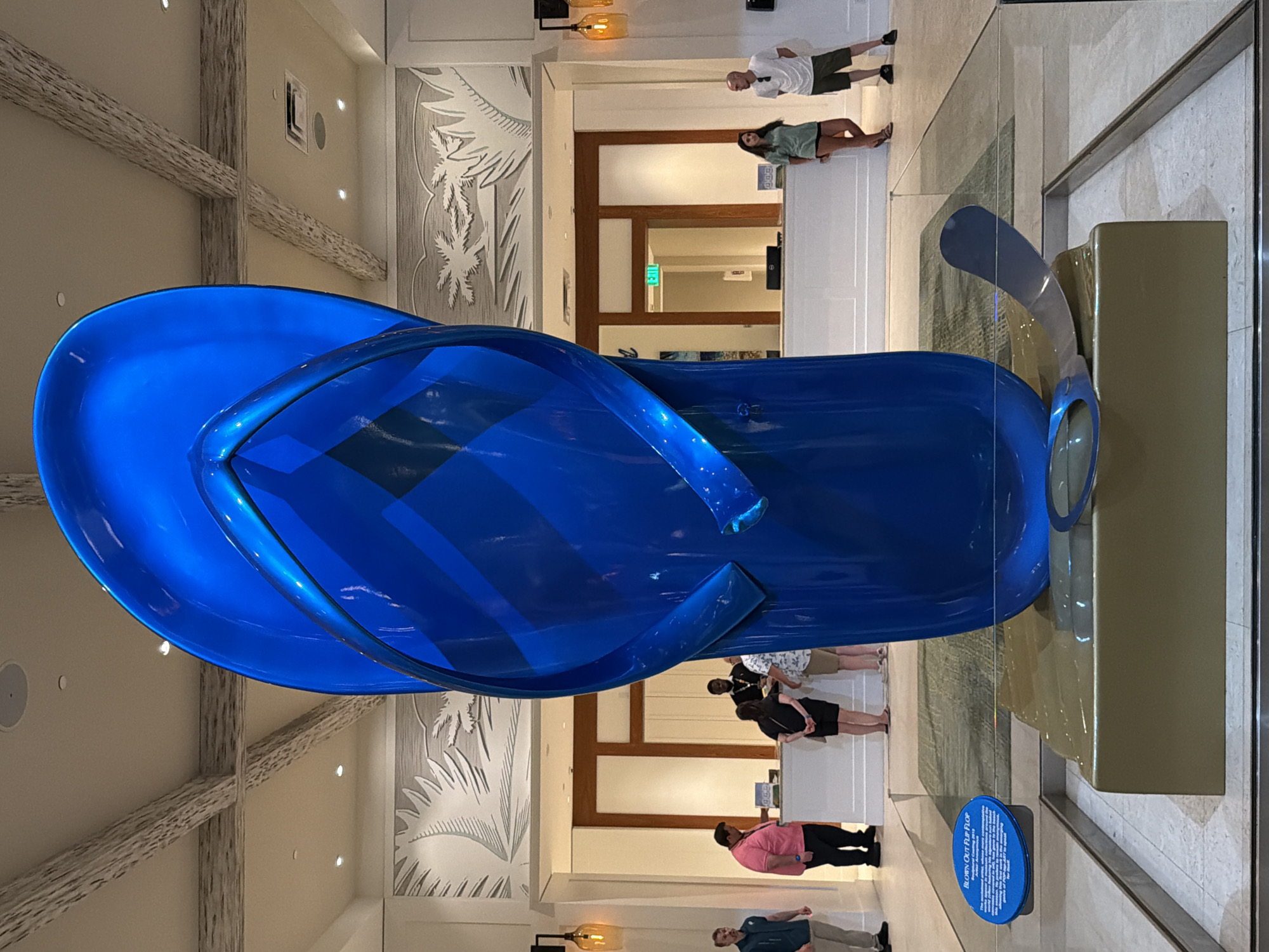 The image depicts a large, vibrant blue sculpture of a flip-flop, likely situated indoors, possibly in a hotel lobby or gallery. The sculpture is polished and reflective, casting subtle highlights. It rests on a golden pedestal, which in turn is placed on a patterned rug. A portion of a metal bottle cap is visible bent beneath the flip-flop sculpture.
To the left of the sculpture, a blue informational plaque with text is visible, partially obscured by the flip-flop. In the background, a reception desk with several individuals is present. One person in a pink shirt is standing behind the desk, and another, a man in a black shirt, appears to be a staff member. Several other individuals are seen interacting at the desk or standing nearby.
Above the reception desk, a decorative wall panel featuring stylized palm fronds and a beach scene is mounted. The ceiling features exposed, light-colored beams. Lighting fixtures are visible, including lamps with orange glass shades to the left and right of the reception area. An "EXIT" sign with green lettering is visible above a doorway in the background.
The overall environment suggests a well-lit, contemporary indoor space. Based on the style of the sculpture and decor, it could be located in a resort or a public area associated with leisure and vacation, potentially in a city like Hollywood, United States of America, known for its entertainment and tourist attractions.