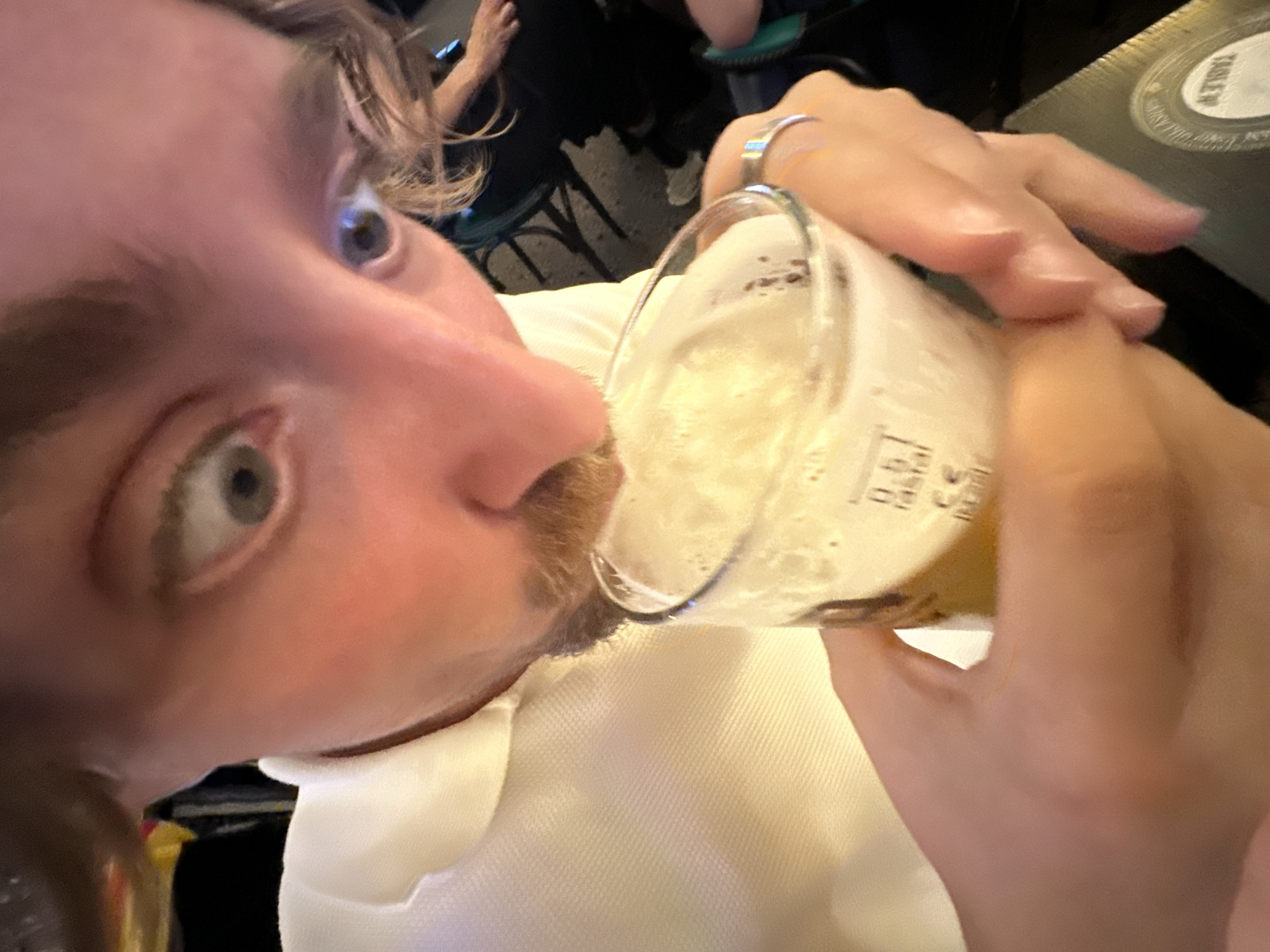 The image shows a close-up of a person with a surprised expression as they drink from a glass of beer. The person is wearing a white polo shirt. The background is dark and appears to be an indoor setting, possibly a bar or restaurant. A small portion of a menu or coaster with the word "TABLE" is visible in the lower right corner. There are no specific landmarks or indicators to pinpoint the location in Belgrade, Serbia.