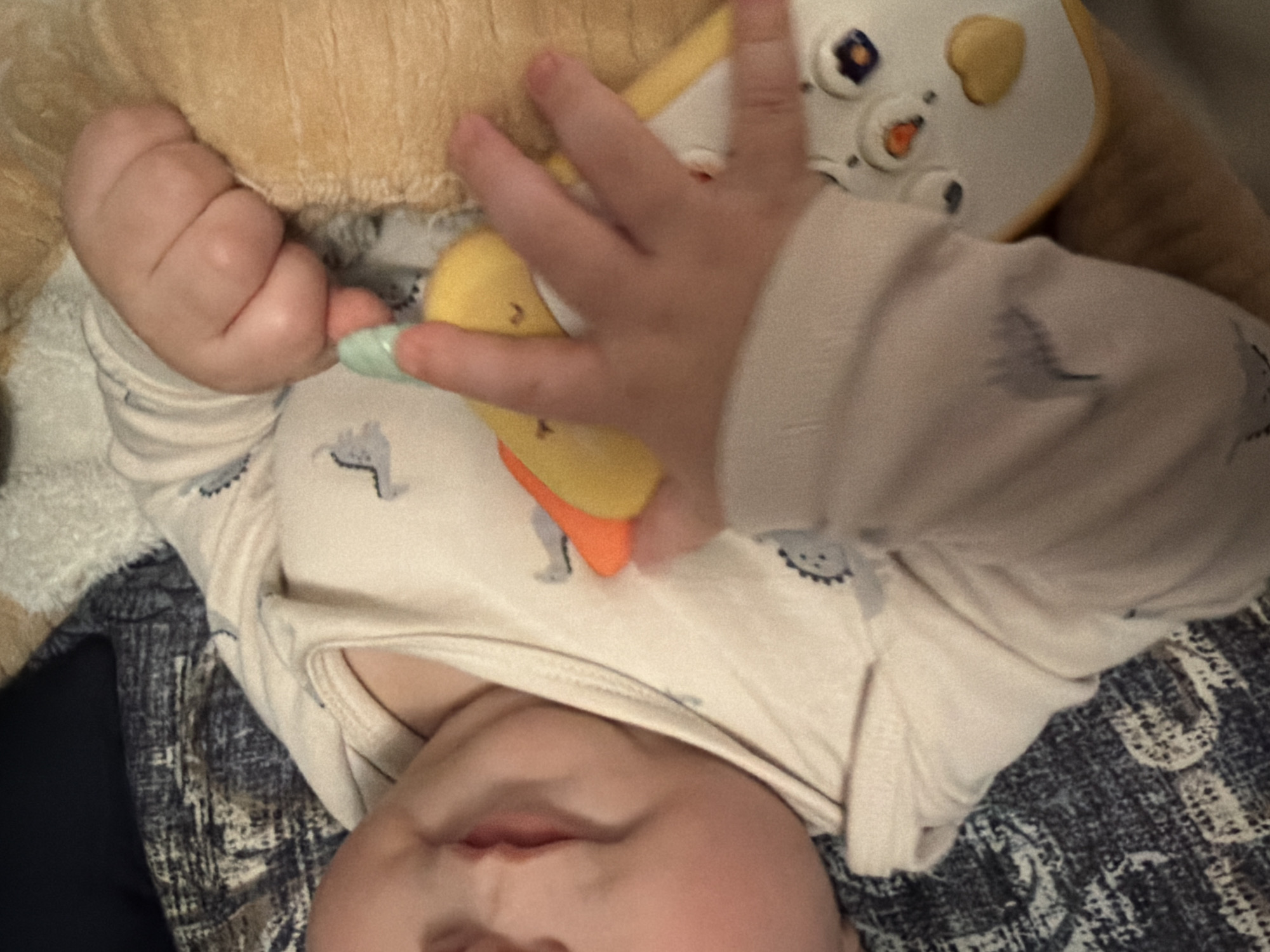 A baby lies on a dark blue and gray patterned surface. The baby is wearing a light-colored onesie with small designs of elephants on it. The baby's face is visible, including closed eyes and mouth. The baby is holding a toy that is partly yellow and orange. Another hand, presumably the baby's, is clenched in a fist. A yellow stuffed animal is nearby. It is not possible to know the location is Yerevan, Armenia, from the image.