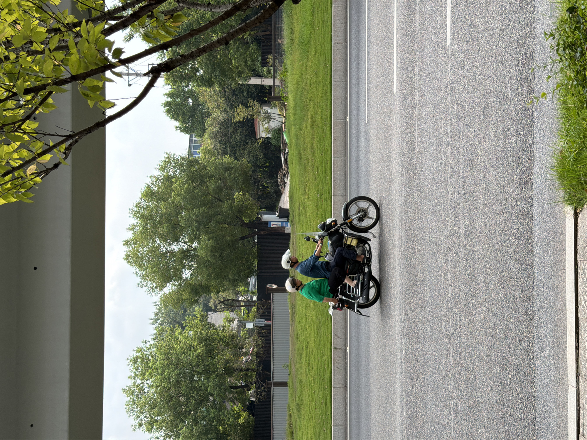 Two people are riding on a motorcycle on a paved road. The person in front is wearing a blue shirt and a white helmet, and the person behind is wearing a green shirt and a white helmet. They are both wearing helmets. The motorcycle is black and has a windshield. The road has white lane markings. In the background, there is a green grassy area, trees, and a fence. There is also a building in the distance. The sky is cloudy. A light-colored concrete structure is visible in the foreground, partially obscuring the view of the road.