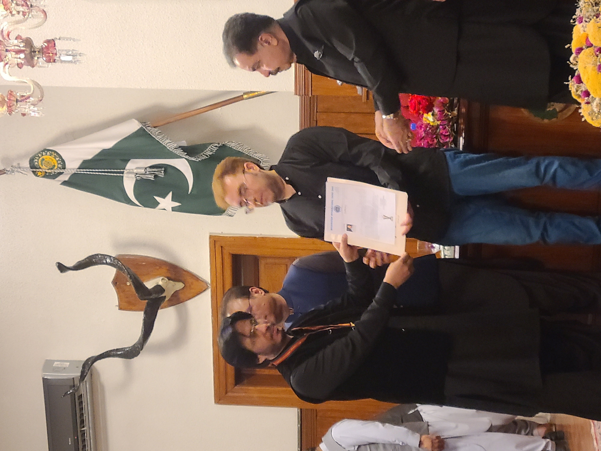 The image depicts a formal indoor gathering in Quetta City Tehsil, Pakistan, featuring several men. The central activity involves the exchange or presentation of official documents. A man with light brown hair and glasses, dressed in a black shirt and blue jeans, holds a stack of papers, one of which prominently displays "INTERNATIONAL" and "FORUM" at the top, alongside visible logos. He appears to be receiving or presenting a certificate or award. Another man, wearing a black shalwar kameez, leans over to observe the documents. To their left, two more men, also in black shalwar kameez, are present, one actively looking at the papers.
In the background, a Pakistani flag hangs vertically, featuring its distinctive green and white design with the crescent and star, and an official emblem near the top. The room is decorated with an ornate chandelier partially visible in the upper left corner. On a wall, a large pair of dark, curved horns are mounted on a wooden plaque, with an air conditioner unit situated beneath them. Vibrant floral arrangements, featuring red, purple, and yellow blossoms, are visible on a surface to the right. The setting suggests an official ceremony or recognition event, with participants exhibiting focused and respectful expressions.