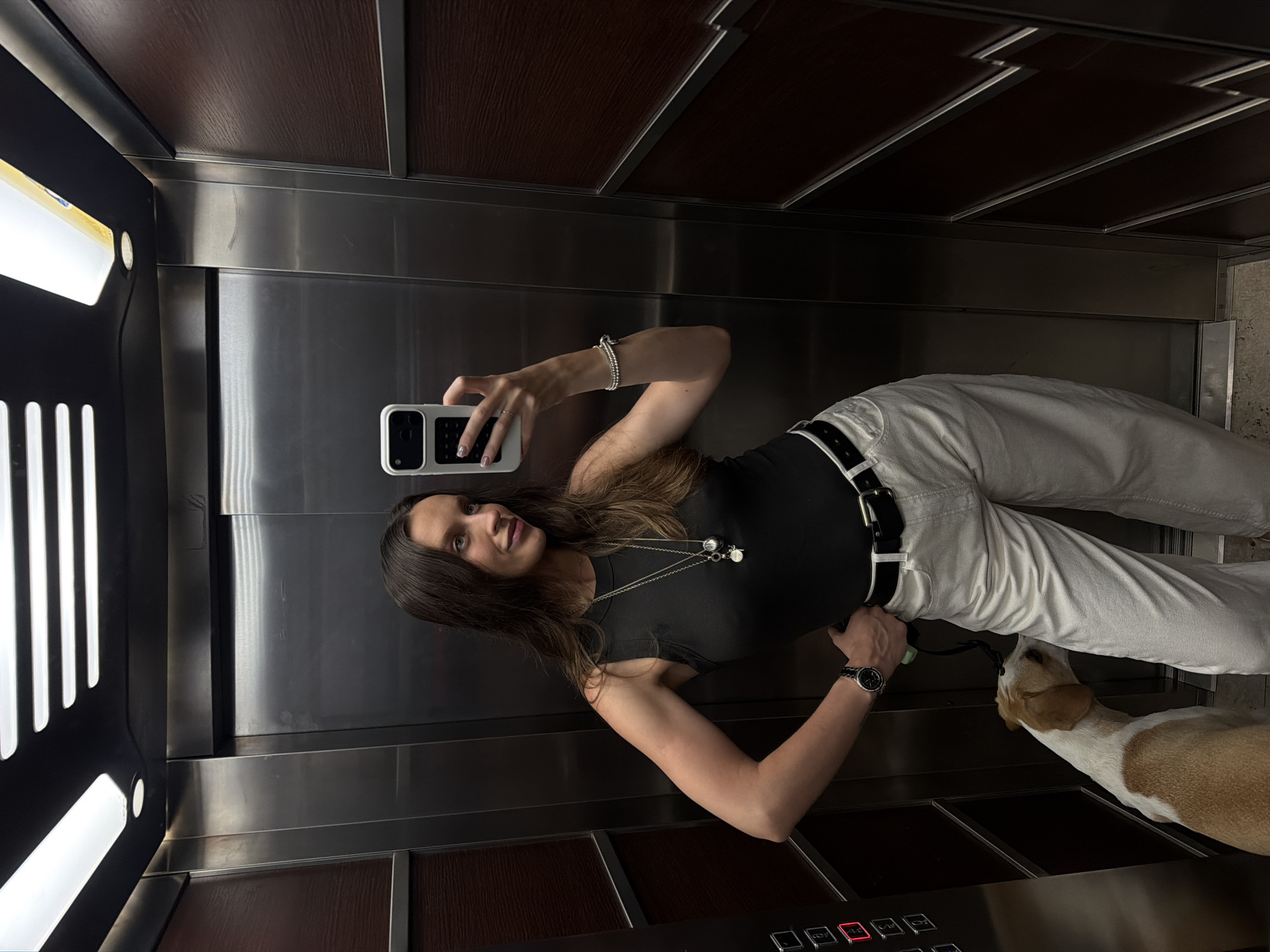 A woman takes a mirror selfie in an elevator in Budva, Montenegro, with a dog. She is wearing a black tank top, white high-waisted pants, a black belt, and a watch. She has long, dark hair and is wearing necklaces and a bracelet. The dog, a medium-sized breed with white and tan fur, is standing next to her and sniffing her pants. The elevator has stainless steel walls and dark wood paneling. The woman is holding a white smartphone in her right hand.