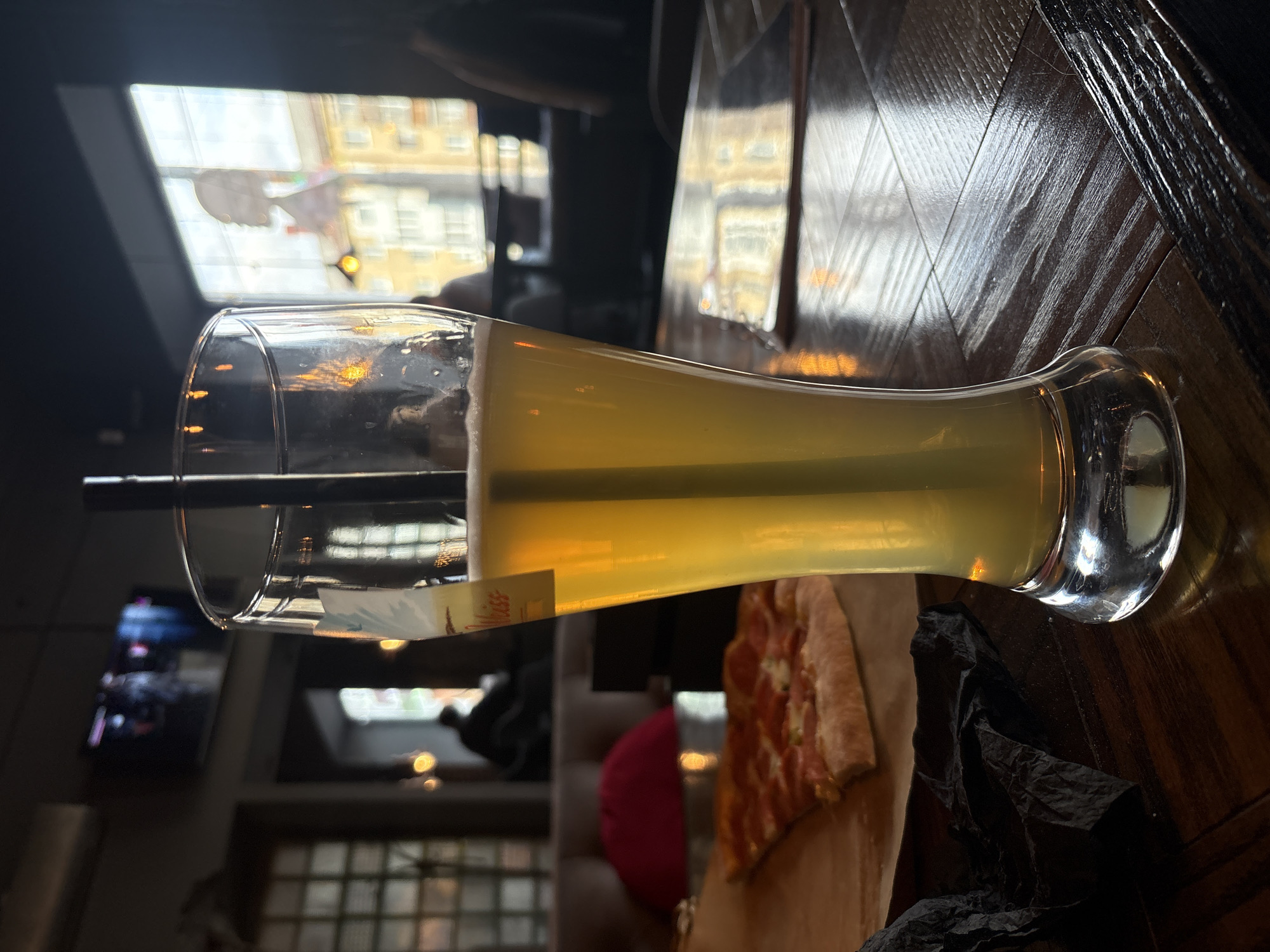 The image depicts a glass of pale yellow liquid with a black straw, likely a beverage, placed on a dark wooden surface. Adjacent to the glass, a partially visible slice of pizza rests on a sheet of parchment paper. The surface appears to be a table, given its material and horizontal orientation.
The background features a window through which buildings are visible, suggesting an urban setting. Additionally, there's a muted television screen and a glimpse of an interior space with a red cushion. Considering the combination of elements, the scene likely takes place in a restaurant or bar, possibly in Perm, Russia, based on the prompt.