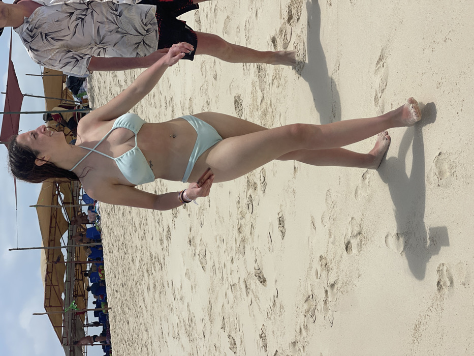 The image displays a sunlit beach scene in Zanzibar, Tanzania. An adult female, with dark, damp hair and a slight smile, walks barefoot on light-colored sand, moving towards the viewer's right. She wears a light blue halter-neck bikini top and matching V-cut bottoms. Visible features include a navel piercing, a small tattoo on her left ribcage, red nail polish on her left hand, and a multi-colored wristband on her left wrist. She holds a small, dark rectangular object between the index and middle fingers of her left hand. To her immediate right, a partially visible adult male walks barefoot, wearing dark swim shorts with a white botanical pattern and a light-colored, short-sleeved collared shirt with a dark leaf pattern. Both individuals cast distinct shadows on the sand. In the mid-ground, several rudimentary beach shelters constructed with light fabric canopies supported by wooden poles are present. Beneath these shelters, multiple indistinct individuals are seated or reclining. The sky above is clear.