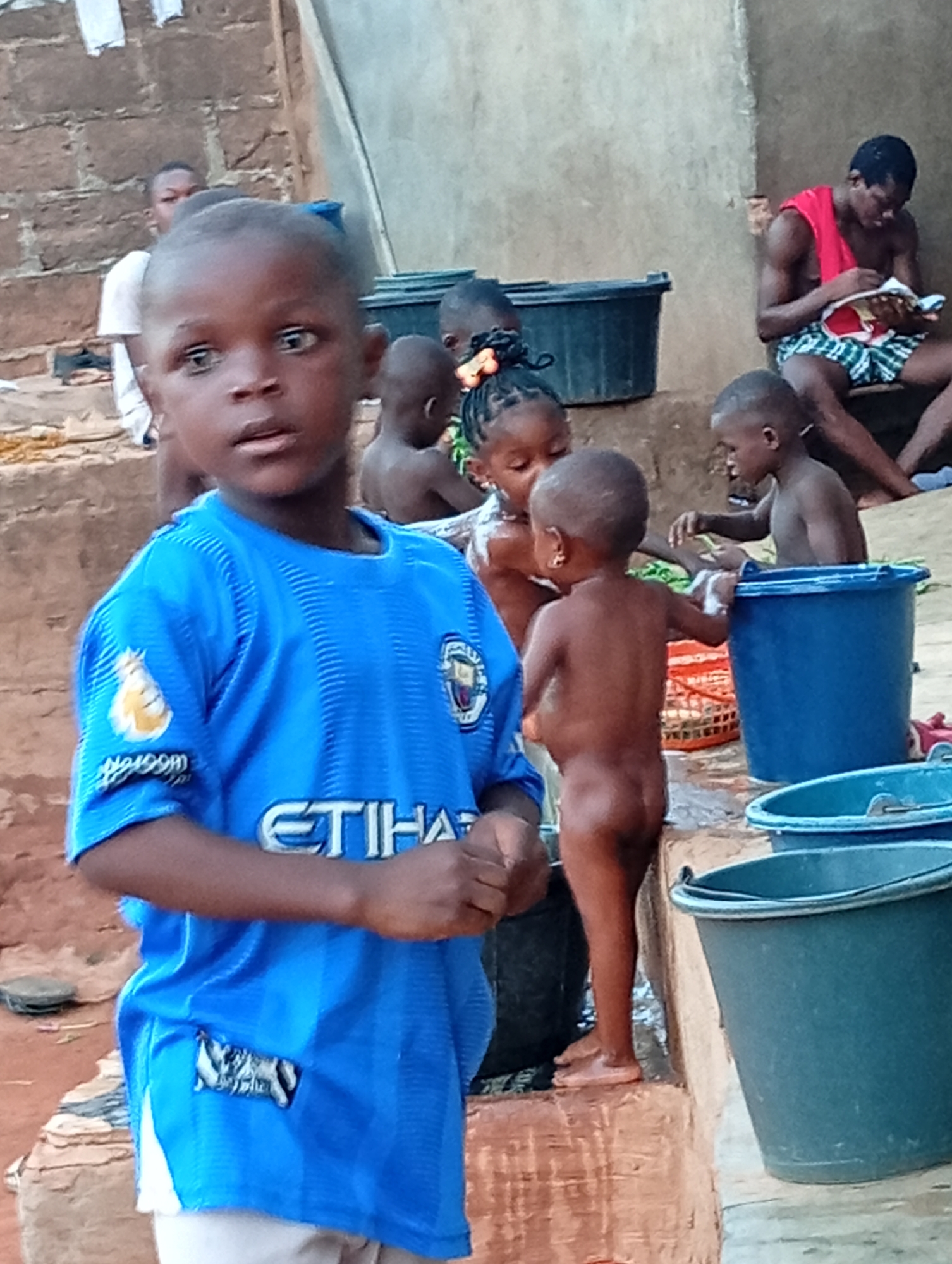 The image shows a group of children and a man in what appears to be an outdoor bathing area in Ogidi, Nigeria. A boy in a blue Manchester City soccer jersey stands in the foreground, looking towards the left. Behind him, several children are bathing using buckets of water. One child is nude. In the background, a man wearing a red shirt and checkered shorts sits and reads. The setting includes a concrete structure and an earthen ground. Clothes hang in the background.