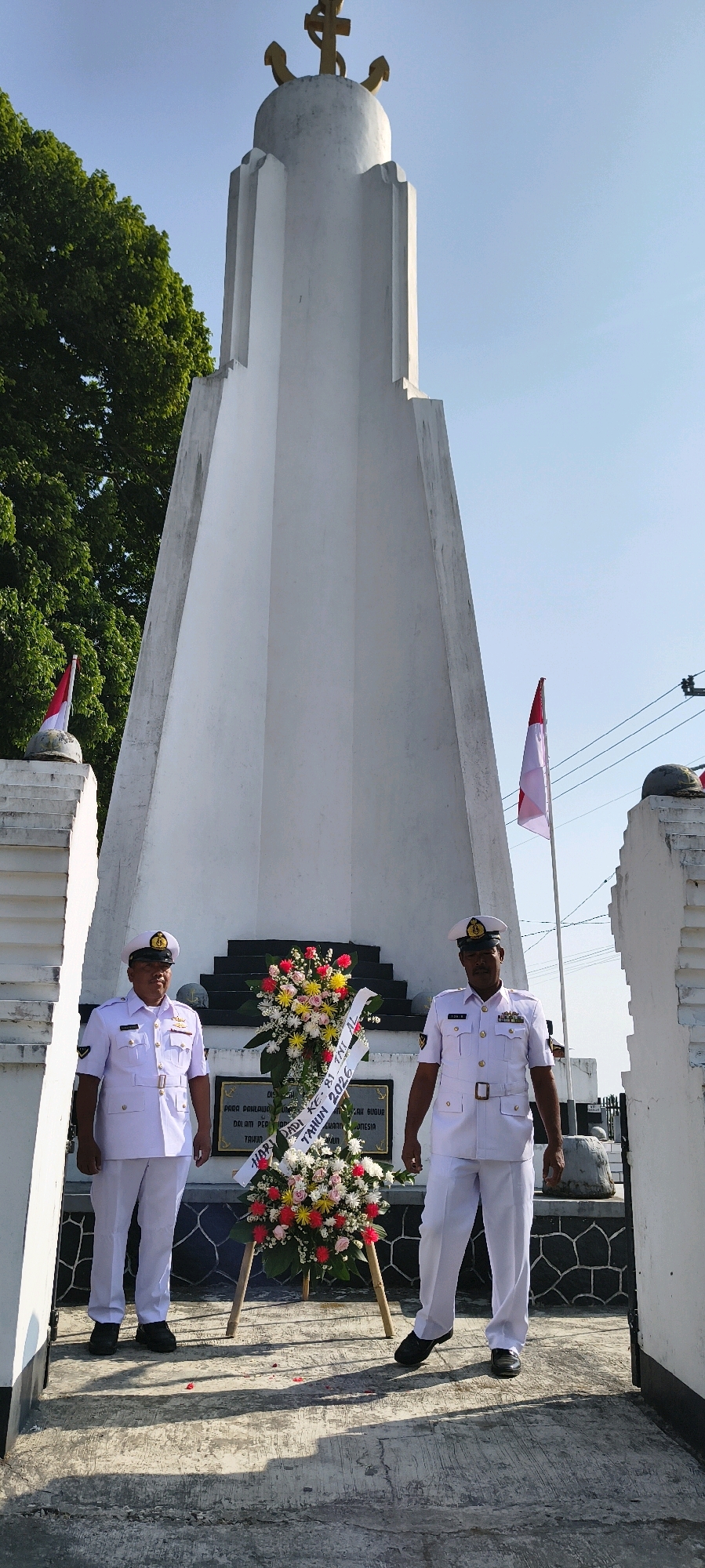 The image captures a ceremonial scene in front of a tall, white obelisk monument. Two men in white naval uniforms stand on either side of a large wreath of colorful flowers adorned with a banner. The banner, partly obscured by flowers, displays text that appears to be in Indonesian, mentioning "Hari Pahlawan" (Heroes' Day) and the year "2026." The monument has a distinct stepped base and a pointed top topped with a golden anchor and cross emblem. Red and white Indonesian flags are visible on smaller pedestals to the left and right of the monument. The environment suggests an outdoor, possibly historic, location with trees and clear sky. The overall tone is formal and respectful, indicative of a commemoration event. The location is identified as the city of Sadamantra, Indonesia, though this specific detail is not visually present in the image itself.