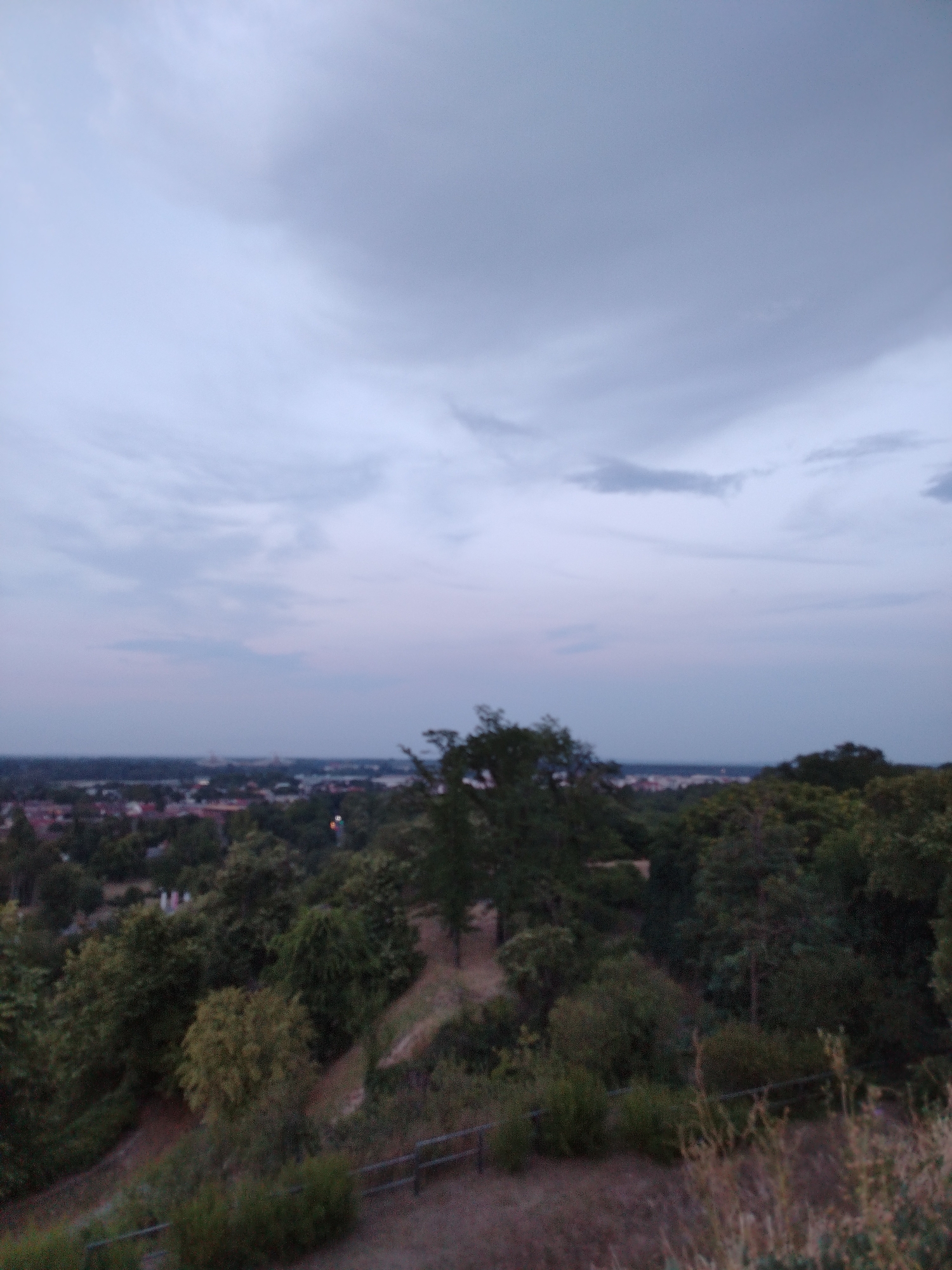 The image depicts an overcast sky over a landscape view from a high vantage point, likely Petrovaradin Fortress in Petrovaradin, Serbia. In the foreground, the terrain is covered with grass and scattered trees. A railing runs horizontally across the lower portion of the frame. Beyond the immediate foreground, a dense collection of trees forms a green canopy. In the middle ground, a city or town is visible, characterized by a mix of buildings with red roofs and scattered vegetation. A faint haze or distance blurs the details of the urban area. The sky is filled with various shades of gray clouds, suggesting either approaching rain or dusk. The overall atmosphere of the image is somber and muted due to the cloudy conditions. There are no visible people or animals in the scene.