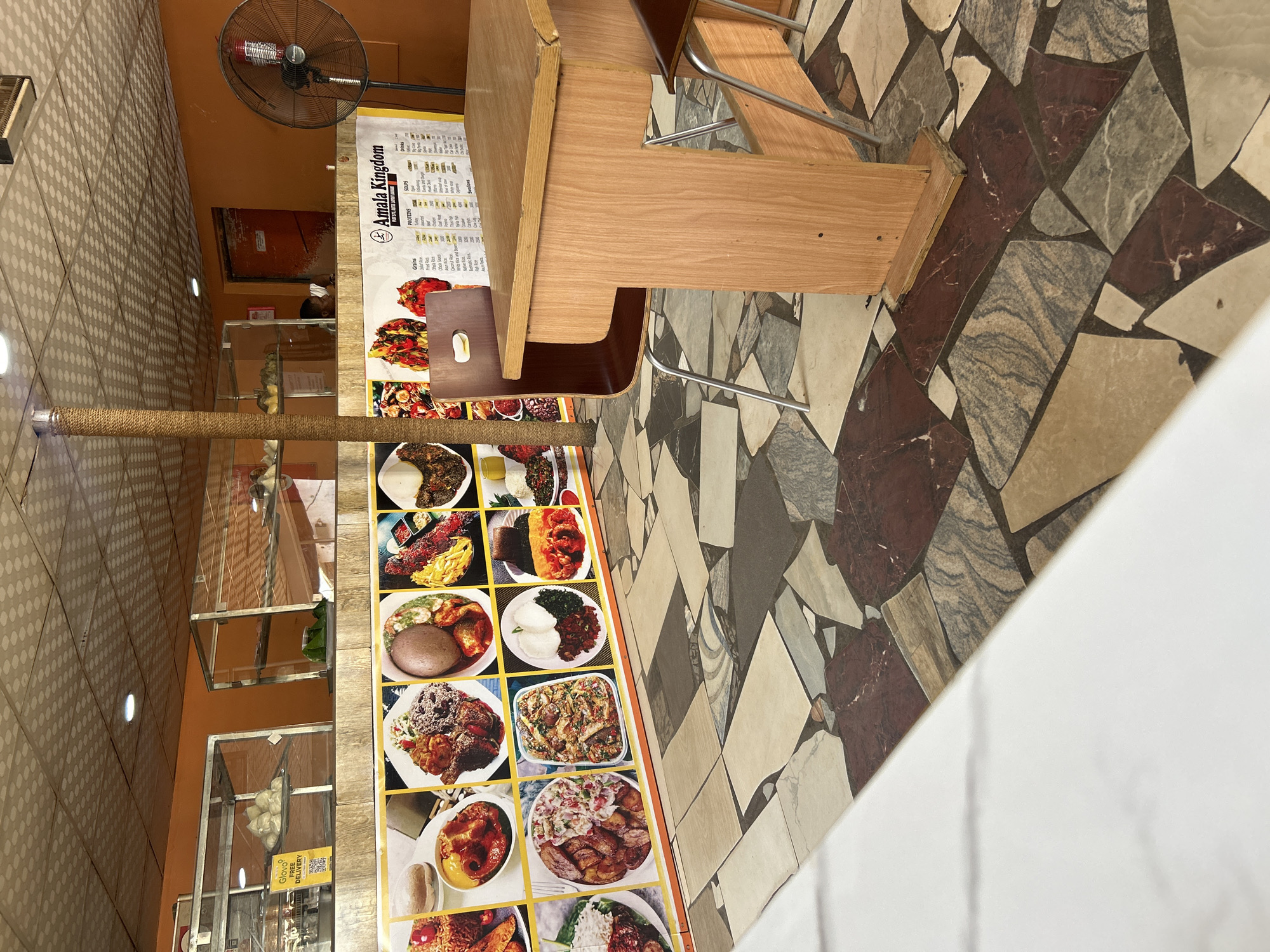 The image shows the interior of a restaurant with an orange painted wall and a patterned tile floor. A wooden table and chairs are set up in the foreground. The wall features a large display of food pictures and a menu board with food items and prices. Display cases with food items are visible behind the counter. A standing fan is positioned in the corner of the room. The overall lighting is artificial and warm.
