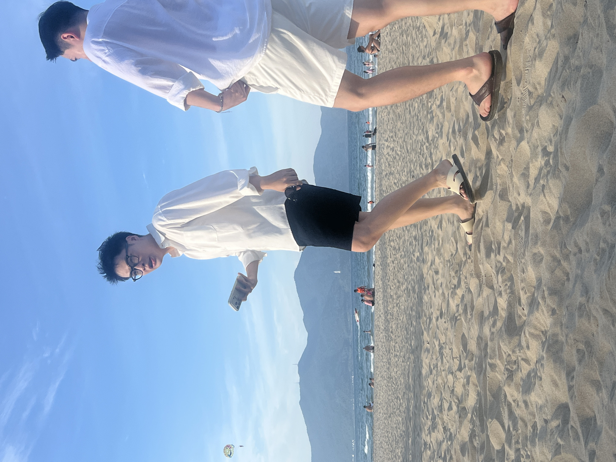 Two young men walk on a sandy beach in Da Nang, Vietnam, with the ocean and mountains in the background. The man on the left, wearing glasses and a white, unbuttoned long-sleeved shirt over a black t-shirt and black shorts, holds a smartphone in his left hand. The man on the right, wearing a white long-sleeved shirt and white shorts, has his hands behind his back. Both men wear sandals. In the distance, people are visible on the beach and in the water. A parasail is visible in the sky. The weather appears to be clear with a blue sky and some clouds. The sun is casting shadows on the sand.