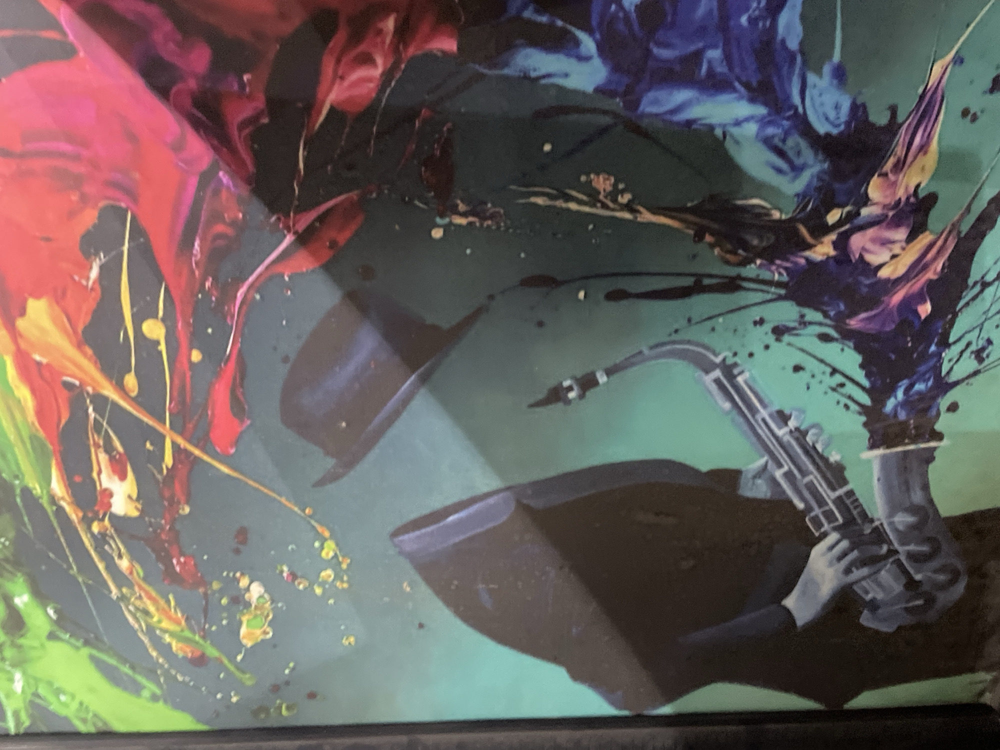 The image is a close-up of a framed art print. The print depicts a person, heavily stylized and shaded in black and white, playing a saxophone. The person is wearing a suit and a fedora, also in black and white. The saxophone is silver. The background is teal, splattered with vivid colors, including red, pink, orange, yellow, green, and blue. The overall effect is an abstract and musical composition. There is no evidence of the location in Abuja, Nigeria.
