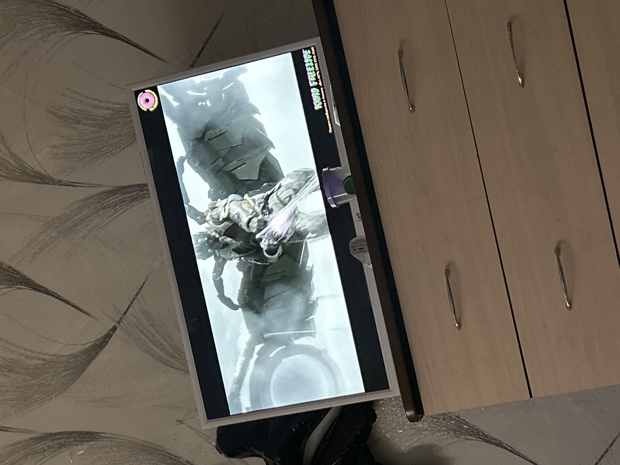 The image, taken in Chaikovsky, Russia, depicts an indoor scene featuring a vertically oriented monitor displaying digital content, positioned next to a light-colored wooden cabinet. The screen is brightly illuminated, showcasing a large, detailed, armored or robotic figure, primarily in shades of gray and green, set against a lighter background. The figure appears to be a character from a video game or animated media.
In the top-left corner of the monitor screen, a distinct pink donut-shaped icon is visible. The top-right corner displays green, stylized text that clearly reads "KWID FREELIVE". Below this, smaller, less distinct text can be partially deciphered as "Правообладатель" (meaning "Copyright Holder" or "Rightsholder") followed by a series of numbers, possibly an identification or phone number, appearing as "ИНН 6776 1950 9109".
The adjacent wooden cabinet is light-toned with vertical lines suggesting multiple doors or drawers, fitted with several elongated, metallic handles. The wall behind the monitor is covered with wallpaper featuring a light background and prominent dark, swirling, feather-like patterns. The scene suggests an indoor environment, likely a room, with artificial lighting. No human subjects are visible.
