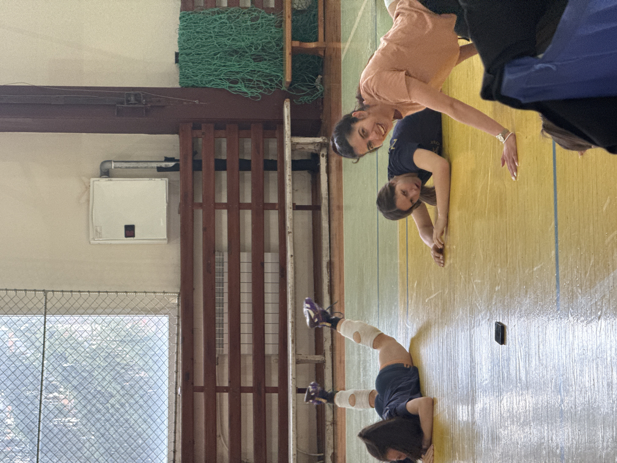 **Location:** The scene appears to be inside a gymnasium or sports hall, possibly in Sofia, Bulgaria, though there is no direct evidence to confirm this.
**People:**
* Two adult women are performing handstands or leaning stretches on the floor.
* A child or young adult female is in a headstand wearing protective padding around the knees.
* Additional unidentifiable people are present in the periphery but partially cropped.
**Objects:**
* A green net is hung on a wall.
* There's a window with a mesh screen.
* A phone is lying on the floor.
* Protective knee pads and athletic shoes worn by one of the individuals.
**Actions and Interactions:**
* Individuals are engaging in athletic stretches or exercises.
* The individuals appear to be smiling and interacting.
**Environment:**
* The room is well-lit.
* The floor is a wooden surface.
* There are wooden structural elements (like support beams or wall partitions) visible.