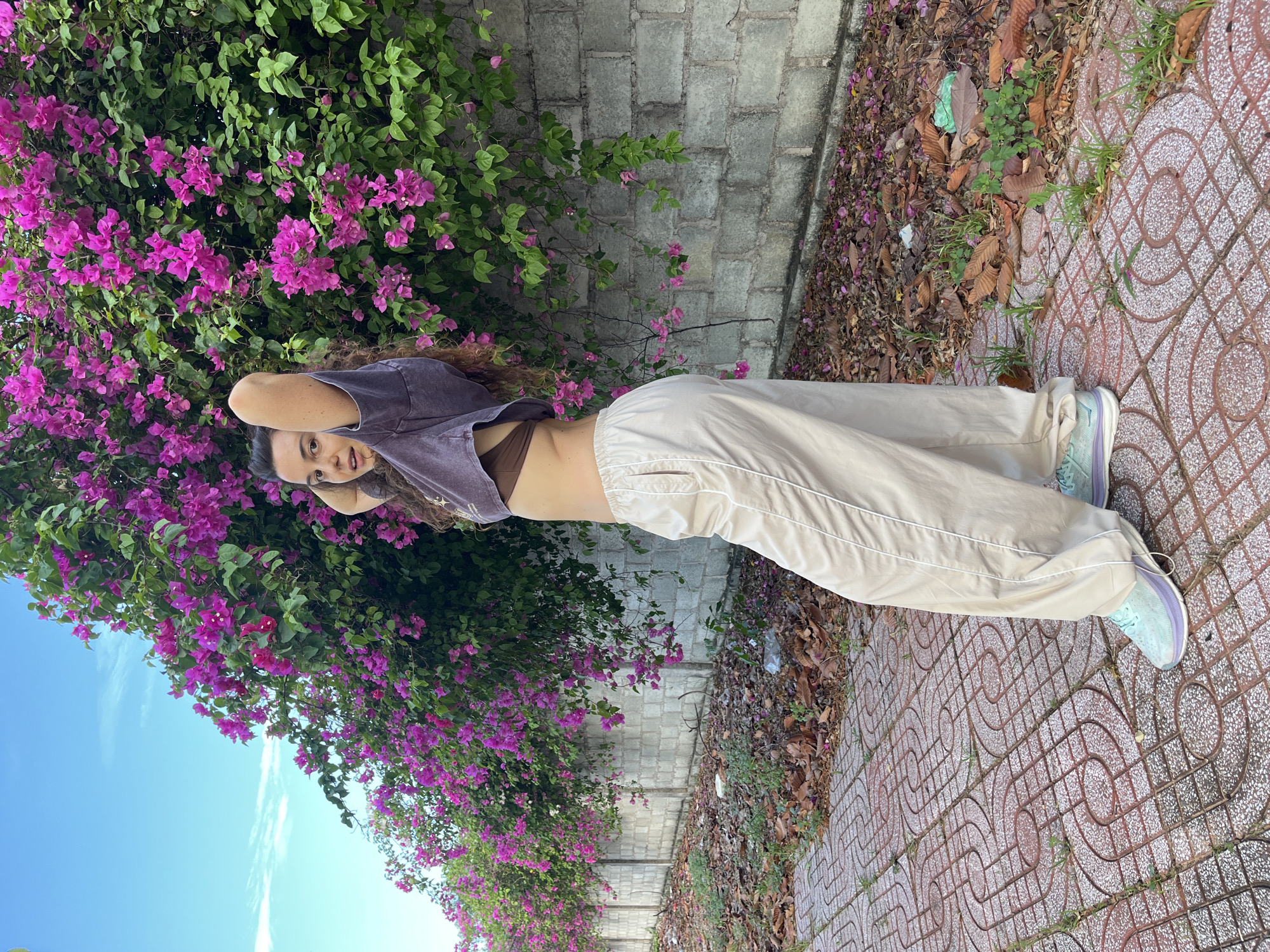 A young woman with curly brown hair stands on a patterned brick pathway next to a wall covered in vibrant pink bougainvillea flowers. She is wearing a brown bralette, a loose-fitting, cropped, dark grey t-shirt, loose beige pants with white trim, and light blue and purple sneakers. Her hands are behind her head. The background consists of a textured concrete wall and a clear blue sky with wispy white clouds. Fallen leaves and some scattered trash are visible on the ground near the wall. The location is identified as Phu Thuy Ward, Vietnam.