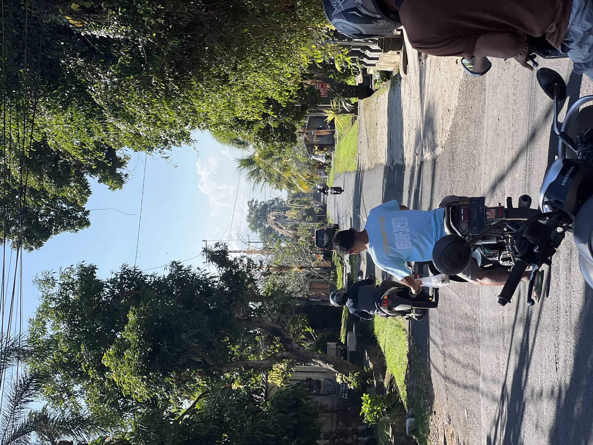 The image depicts a street scene in Sukawati, Indonesia. Two individuals are riding motorcycles on a paved road lined with lush green trees and vegetation. The rider in the foreground is wearing a light blue t-shirt with text that appears to be "BRUIN BORN BRUIN BRED BRUINS TIL THE DAY WE'RE DEAD" and gray shorts. They are holding a white plastic bag in their right hand. The motorcycle has a license plate that reads "DK 5606 BW" and "BALI EXP." The rider behind them is wearing a dark jacket and a helmet. Further down the road, another motorcycle is visible, also with a rider wearing a helmet. A dark SUV is parked on the side of the road in the distance, and several buildings and structures are present on either side of the street. Overhead power lines are strung across the sky, and the sky is a clear blue with scattered clouds. The overall impression is a sunny day in a tropical environment.