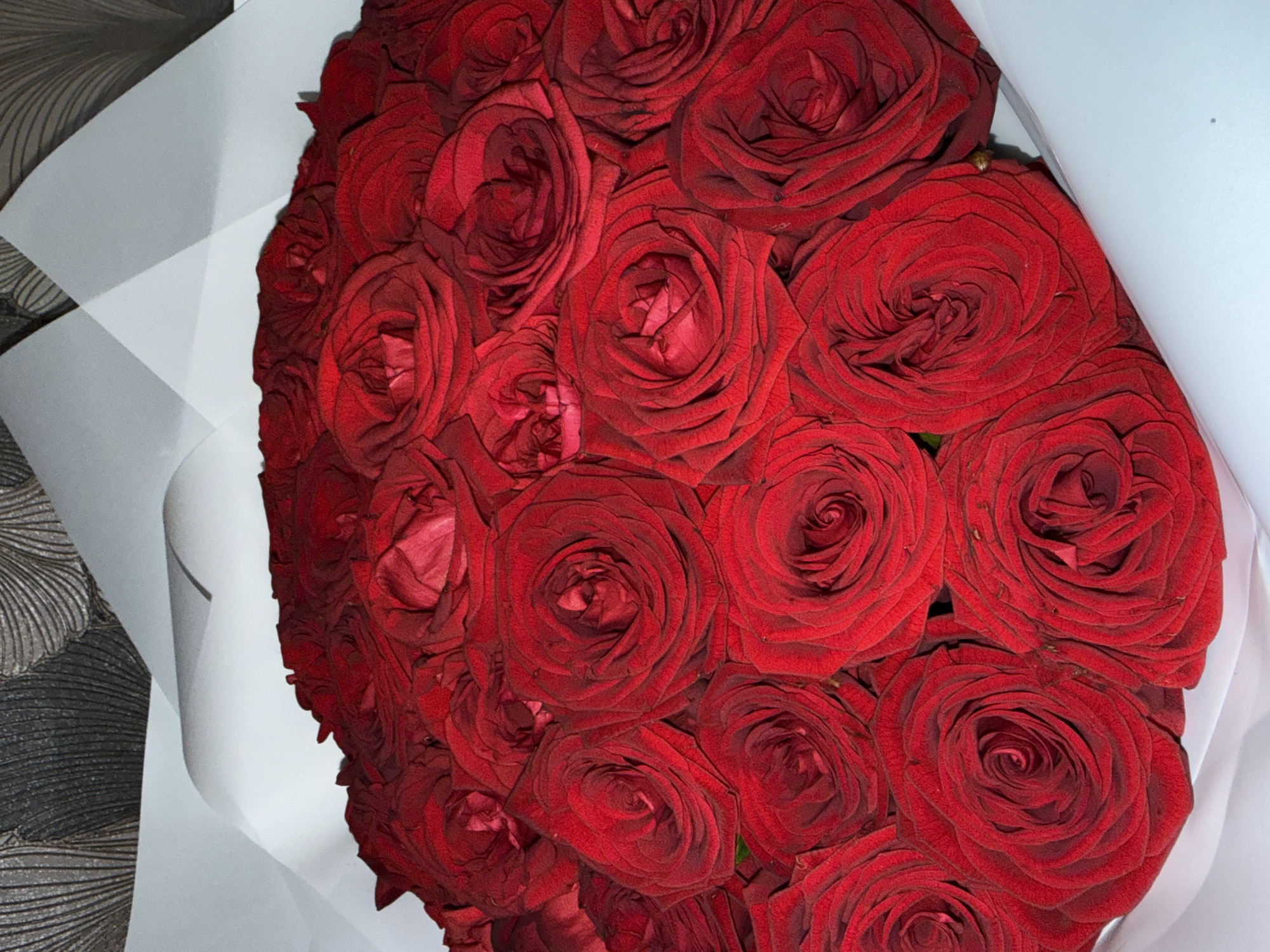 This is a close-up, overhead shot of a large, dense bouquet of deep red roses, presented against a textured background and wrapped in white paper. The bouquet occupies the majority of the frame, positioned towards the center-right.
The roses are tightly packed, showcasing multiple layers of velvety, dark red petals. Their rich color is consistent throughout the bouquet, and individual petals display fine textures and subtle variations in shade. Some petals exhibit minor discoloration or drying at their edges, suggesting a slight aging or a natural characteristic of the rose variety. A hint of green foliage is barely visible amongst the red petals in some sections.
The roses are partially enveloped by crisp white wrapping paper, visible on the left, top, and right edges of the bouquet. The paper features gentle folds and creases, adding depth to the composition. To the far left, behind the white wrapping, a dark, textured surface is visible, possibly a patterned wallpaper or fabric. This background features a repeating, organic or wave-like design in shades of grey and black.
The lighting is soft and even, highlighting the intricate details of the rose petals and the folds in the wrapping paper without casting harsh shadows. No people or visible text are present in the image. The scene depicts a still life, likely indoors, in Izhevsk, Russia.