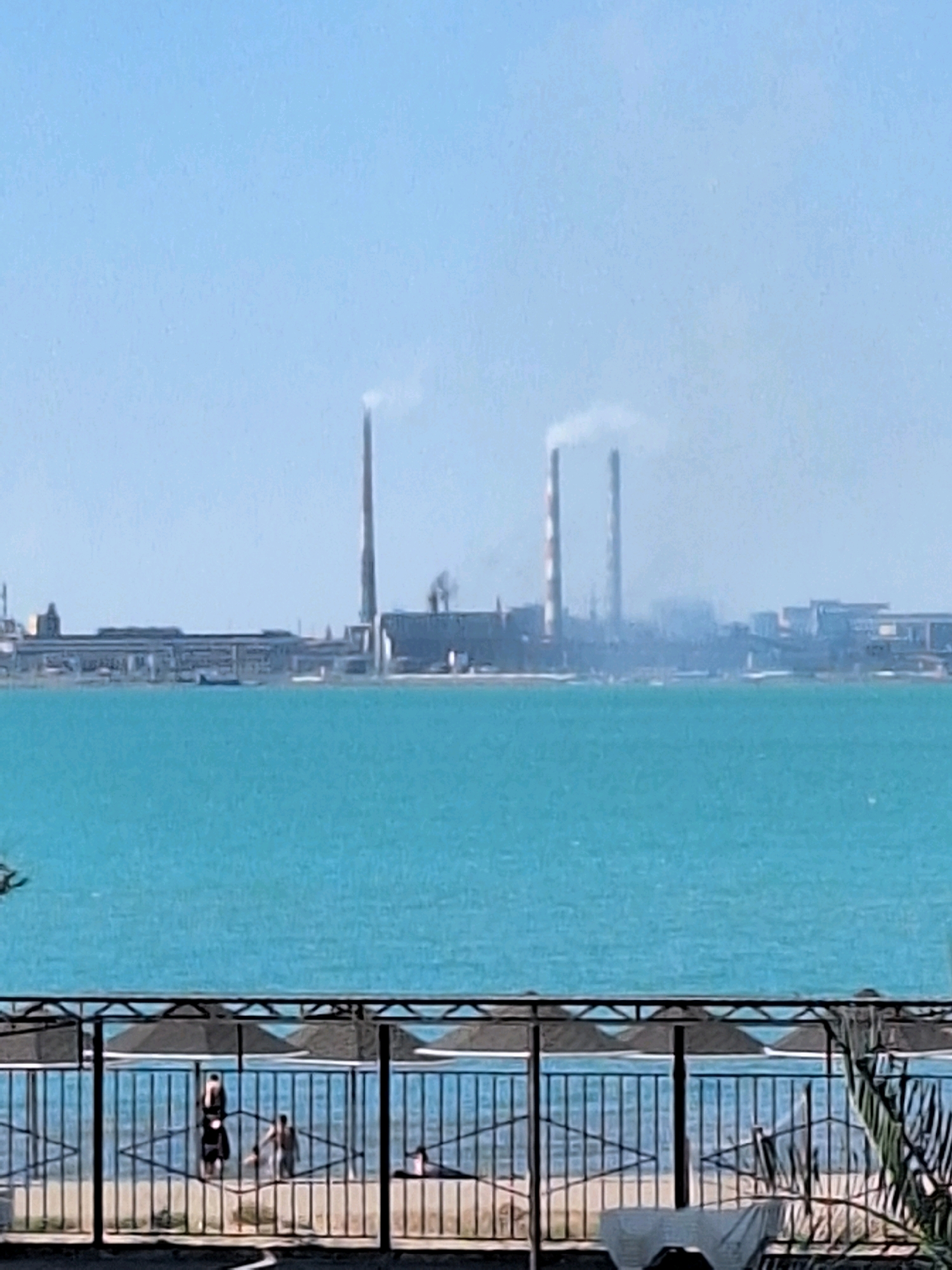 The image depicts a wide-angle view of the Balkhash Lake in Balkhash, Kazakhstan. In the distance, a large industrial complex is visible, with several tall smokestacks emitting plumes of white smoke into the sky. The industrial structures are a mix of modern and older buildings, suggesting a working factory or power plant. The lake's water is a vibrant turquoise color. In the foreground, a black metal fence runs horizontally across the lower portion of the frame. Behind the fence, several people are present, some of whom appear to be in the shallow water. Beach umbrellas are also visible behind the fence, and a lounge chair is partially visible in the bottom right corner. The sky is a clear, pale blue, indicating a sunny day.